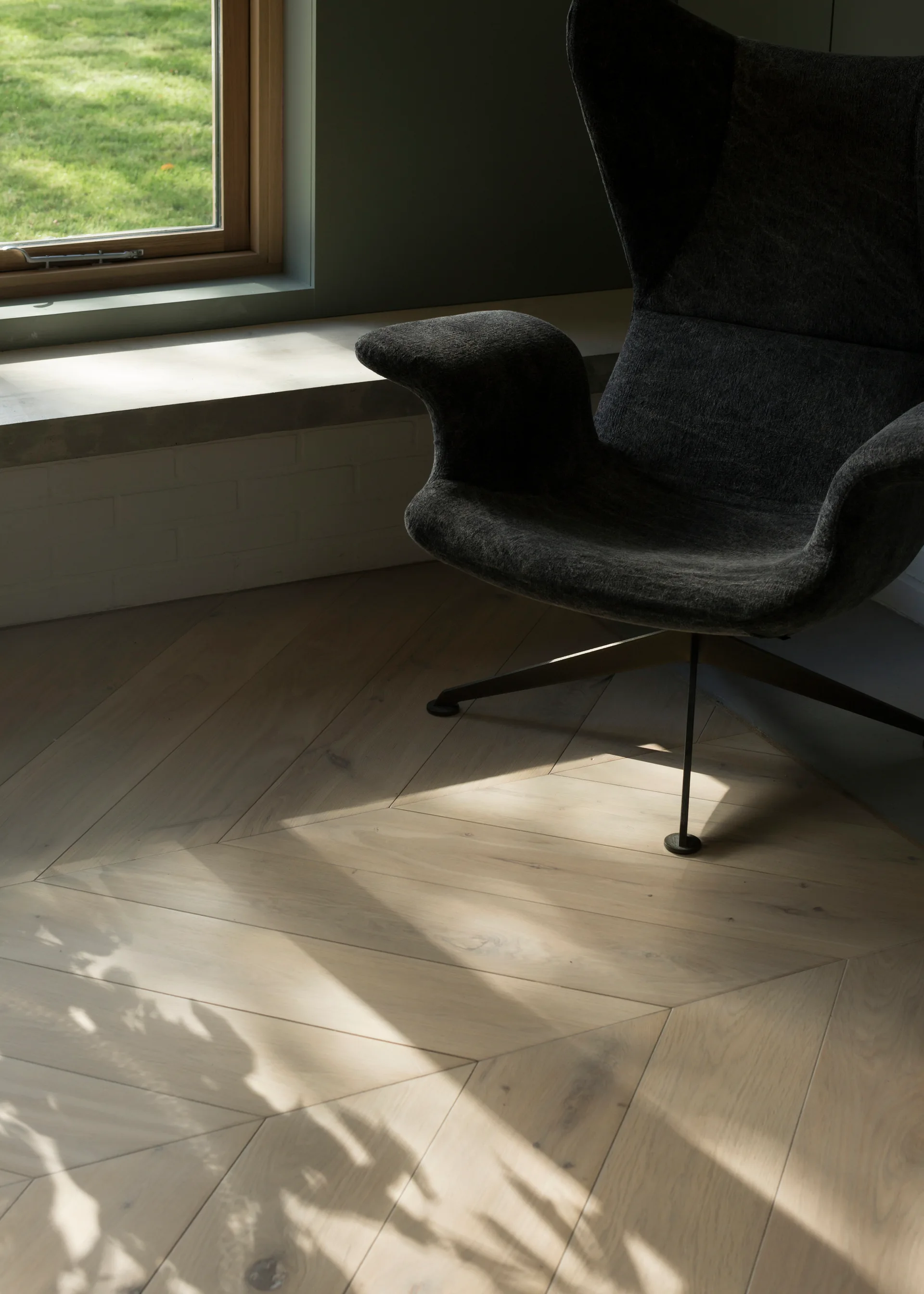 A modern dark grey upholstered armchair stands on a light-toned chevron wood floor near a window.