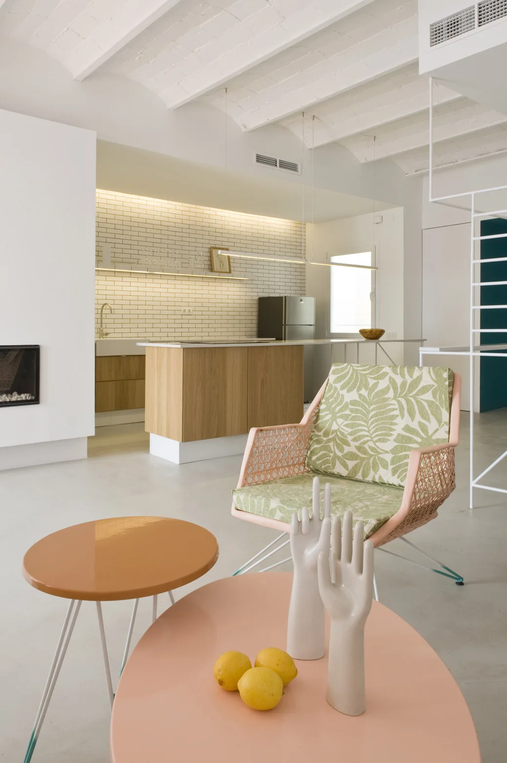 A bright open-plan living room and kitchen features a pink patterned armchair, round pastel coffee tables, and a wood-toned kitchen island.