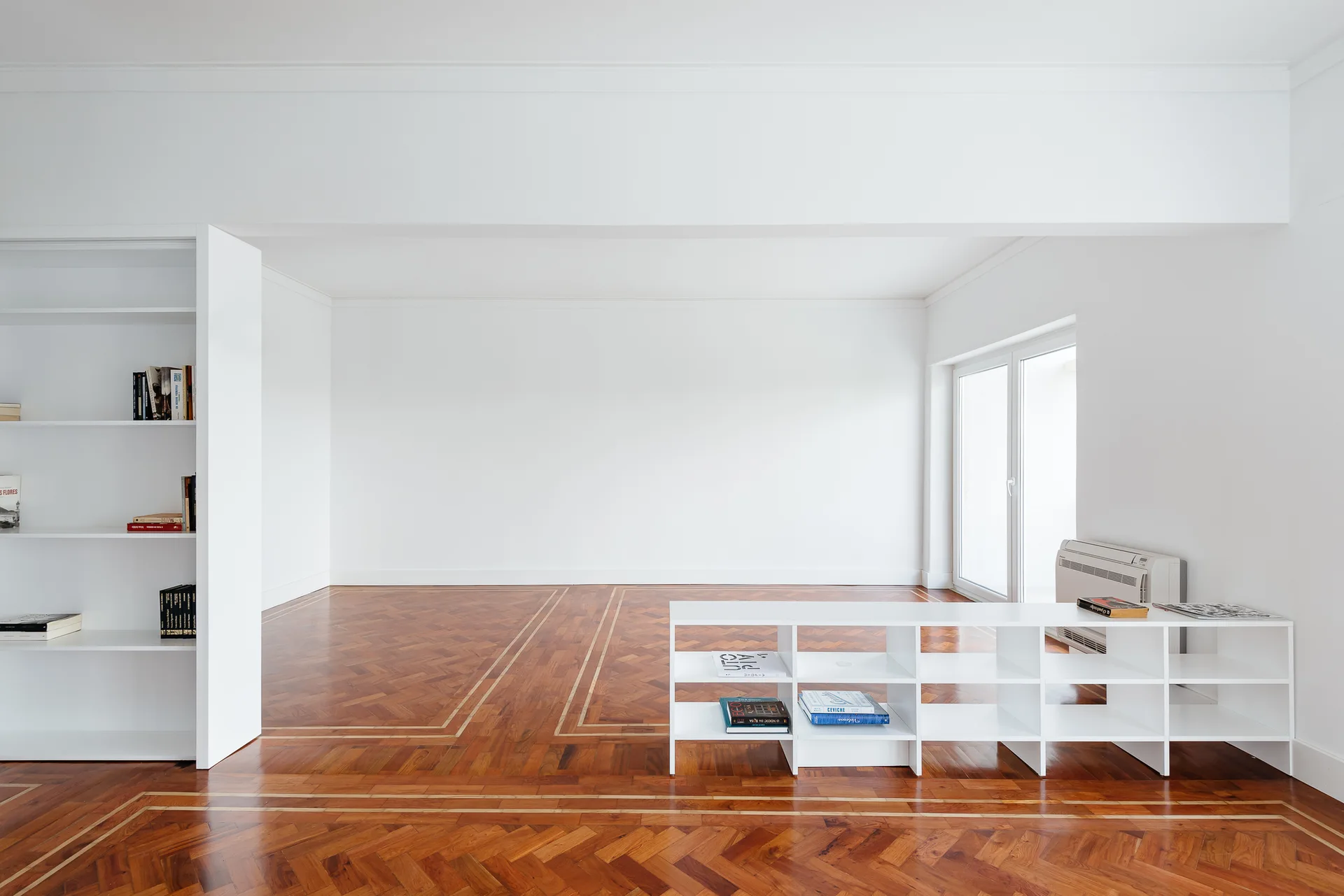A bright white minimalist room features polished brown herringbone wood flooring with decorative inlay borders and built-in white shelving.