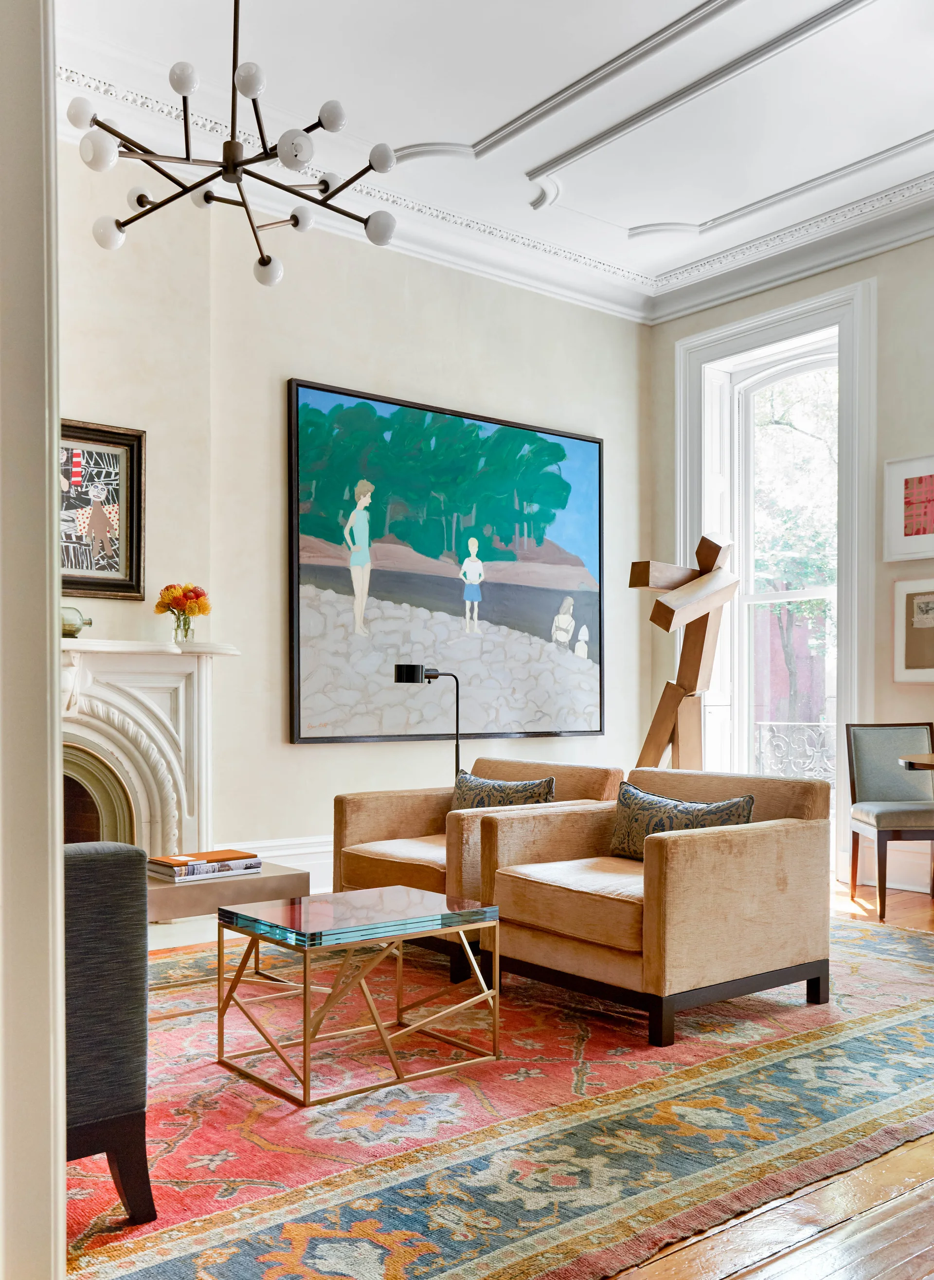 Two beige velvet armchairs sit on a colorful patterned area rug in a living room with cream walls, a fireplace, and a large scenic painting.