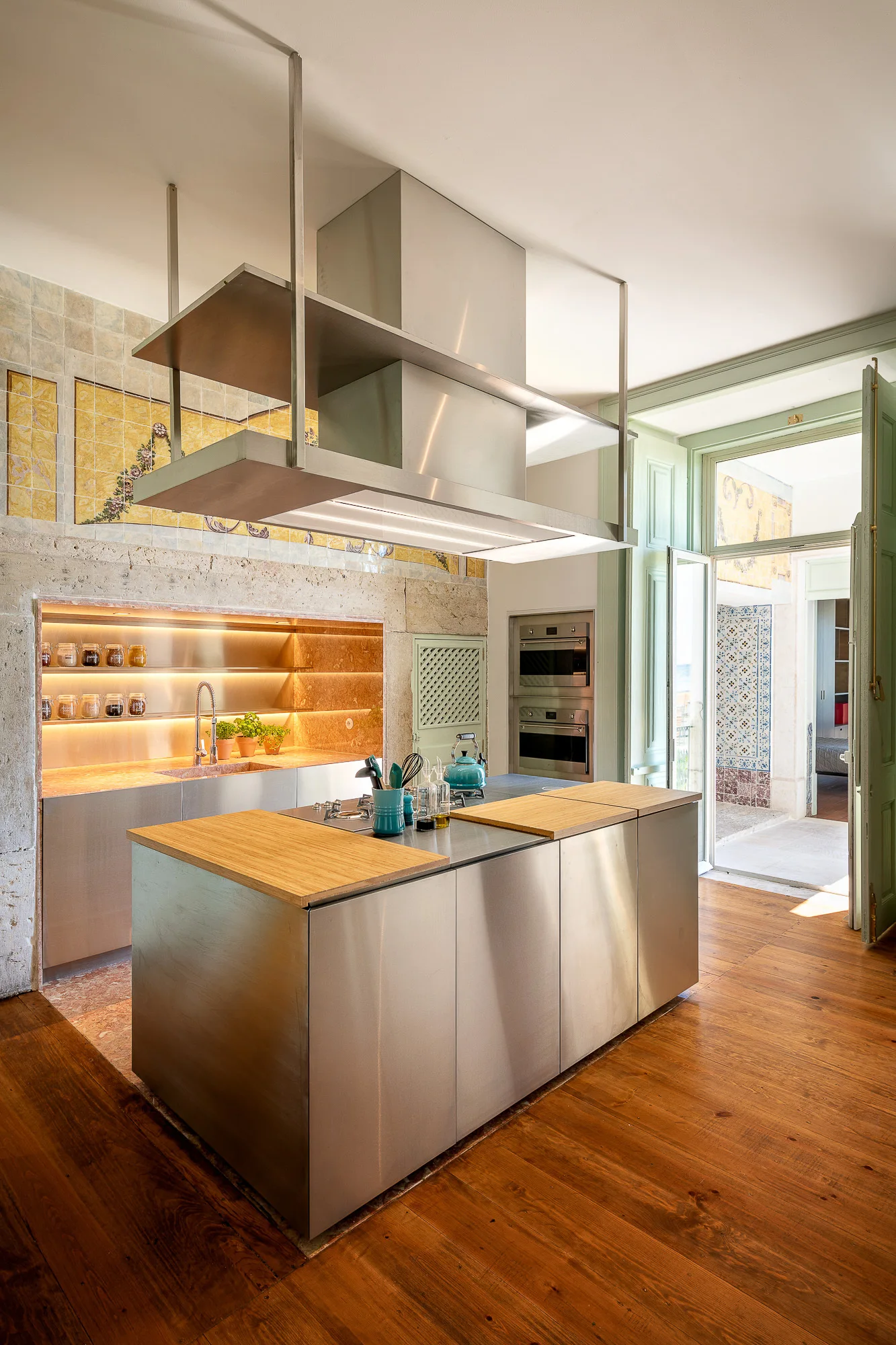 A modern kitchen featuring a stainless steel island with wood inserts, a large suspended hood, wood floors, and a stone alcove with integrated lighting.