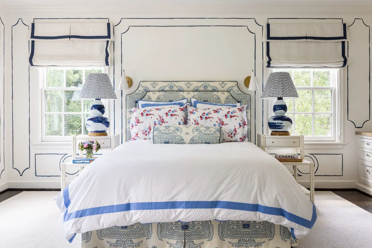 A symmetrical white bedroom features a bed with blue patterned bedding and a headboard, flanked by matching nightstands, lamps, and windows.
