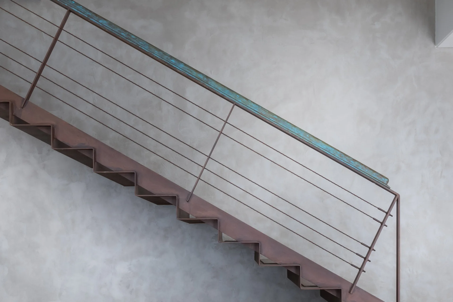 A metal staircase with a rusted finish and a turquoise patina handrail is set against a textured grey plaster wall.