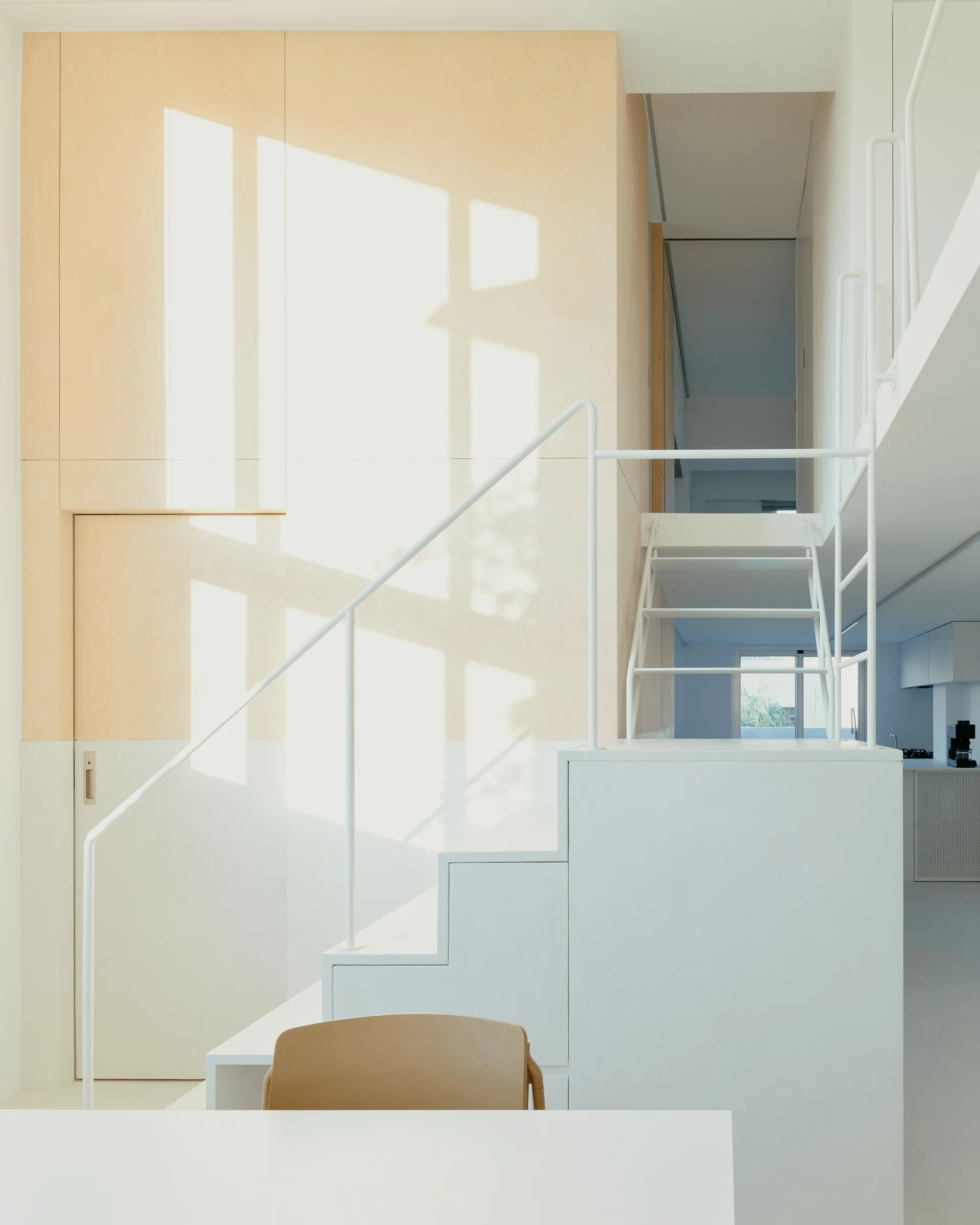 A minimalist white metal staircase stands against a tall accent wall made of light-colored plywood panels with integrated flush doors.