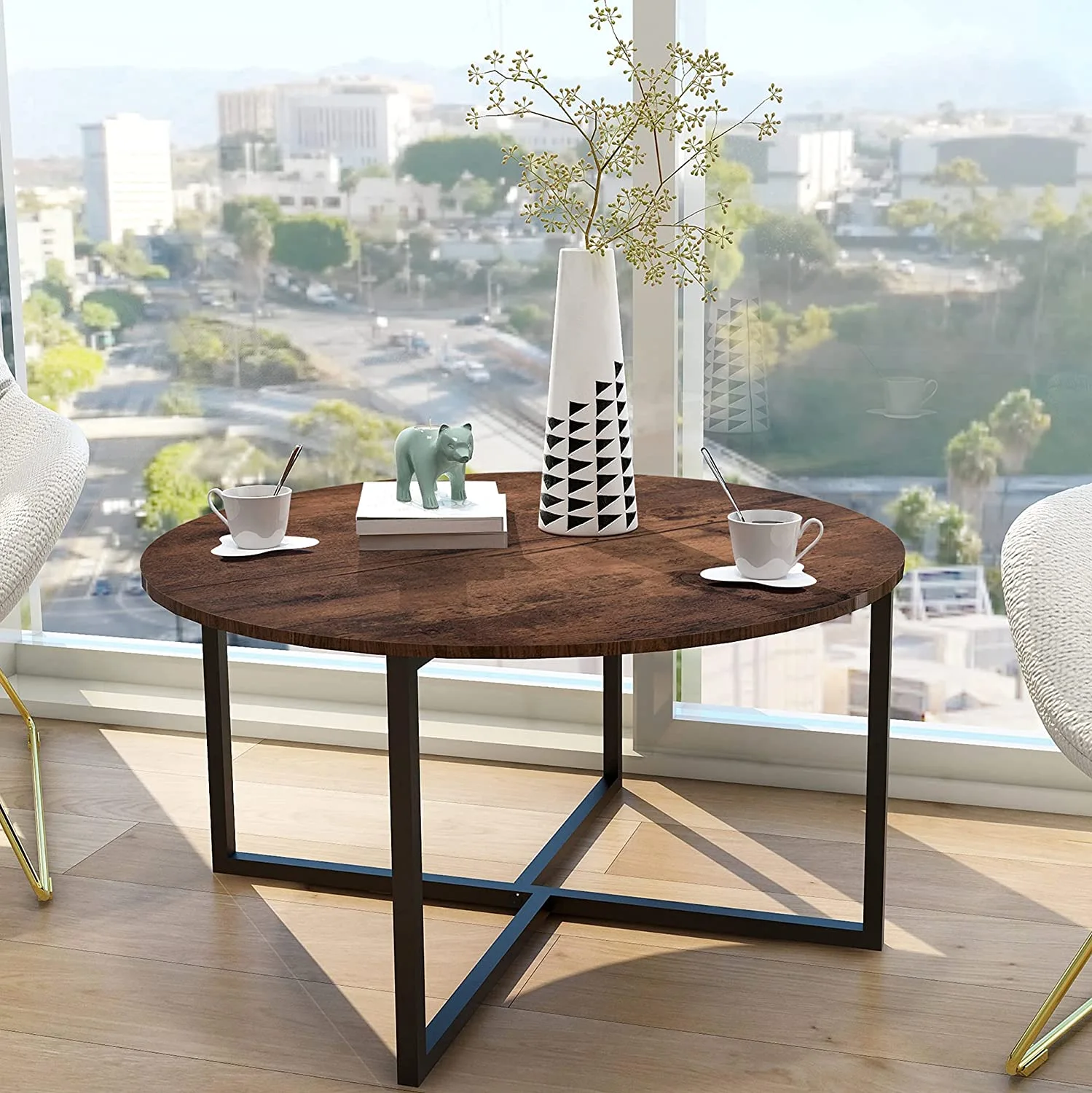 A round wood-topped coffee table with a black metal base sits on light hardwood floors near a large window overlooking a cityscape.
