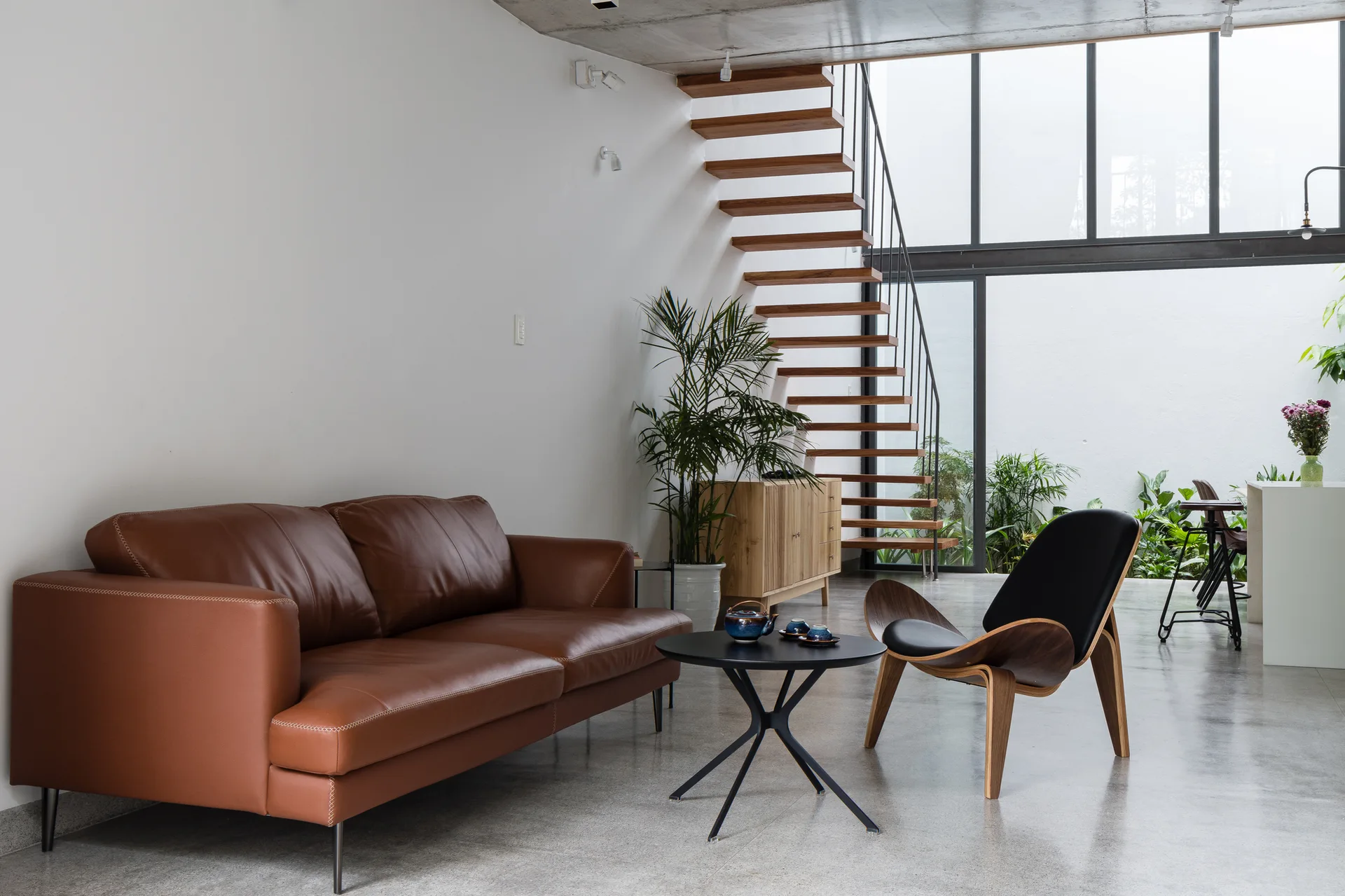 A modern living room features a brown leather sofa, a black curved accent chair, and a floating wooden staircase set against a glass wall.