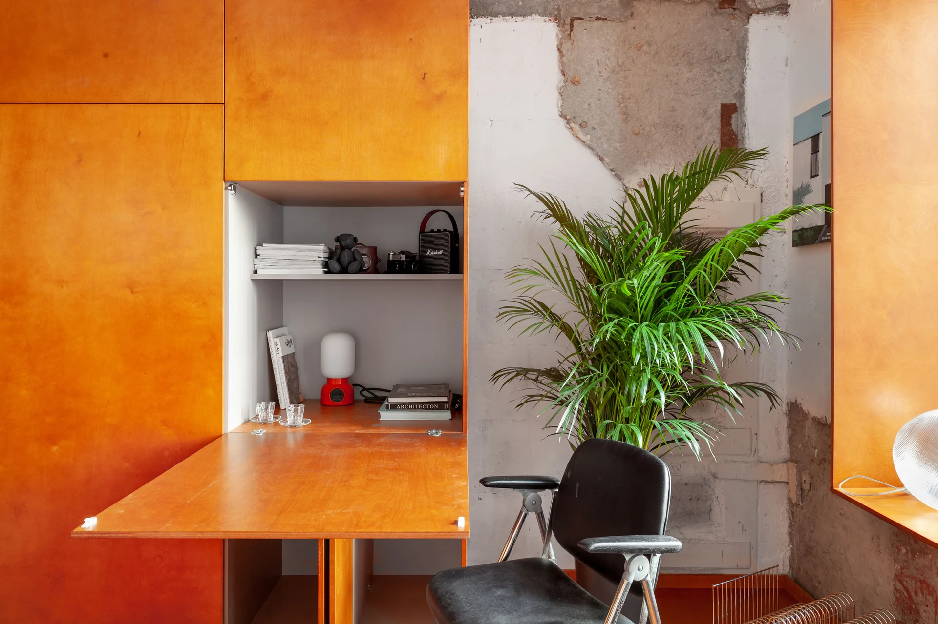 An orange wood built-in unit with a fold-down desk, a black office chair, a green potted palm plant, and an unfinished plaster wall.