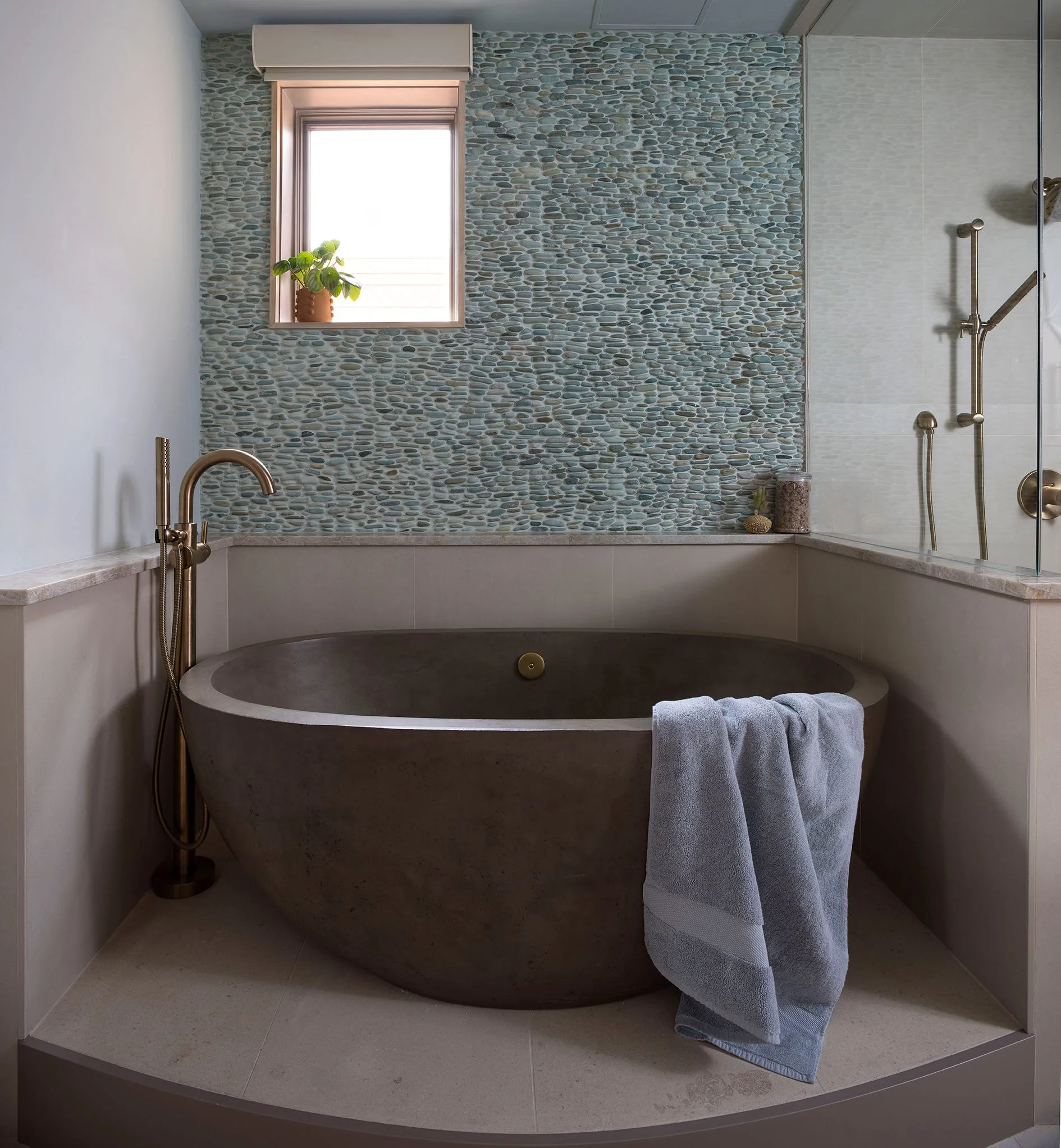 A dark concrete freestanding bathtub sits below a small window in a bathroom featuring a teal-gray pebble tile accent wall.