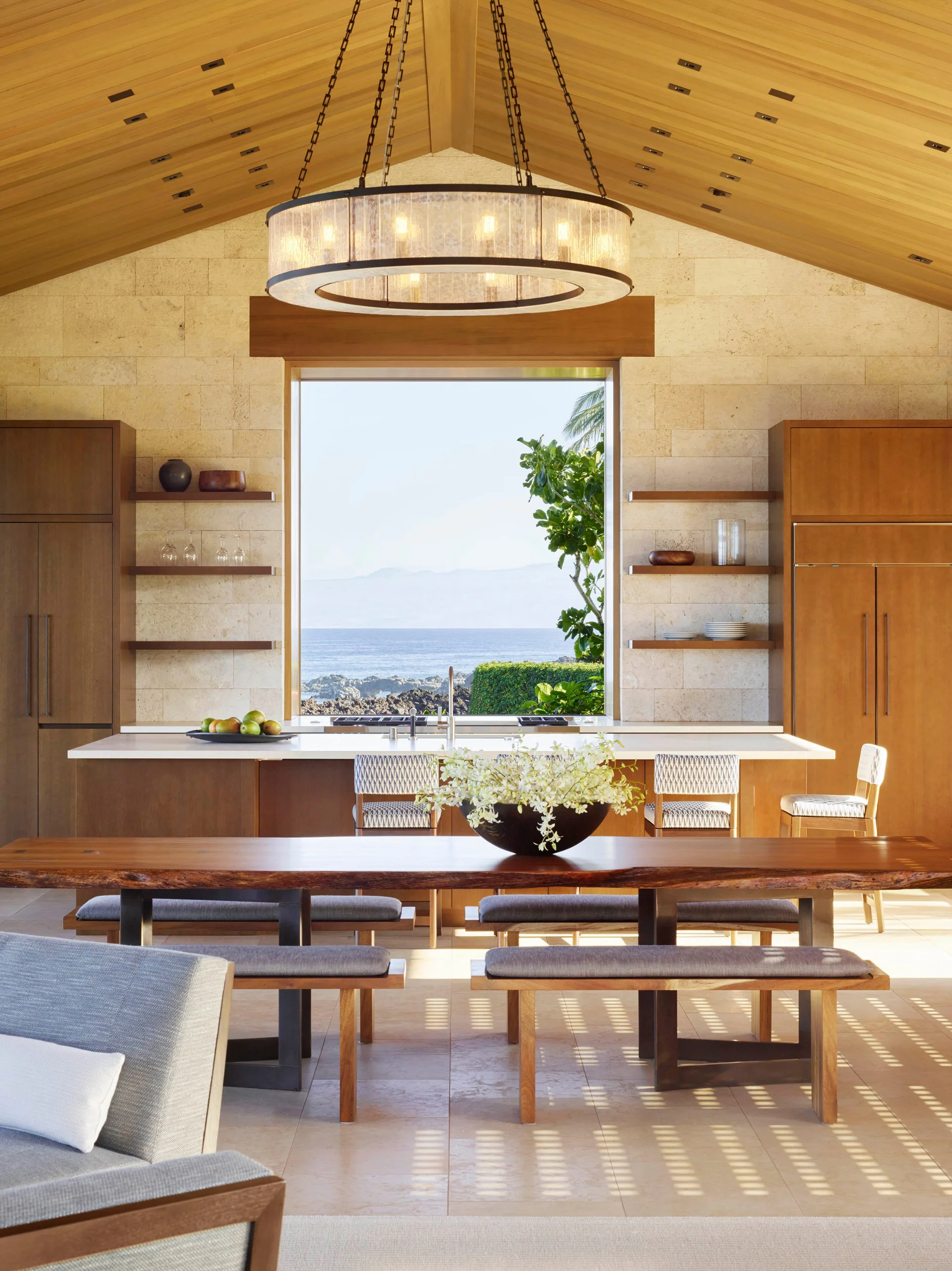 A modern coastal kitchen features a large window with an ocean view, a vaulted wood ceiling, and a central island flanked by built-in wooden shelving.