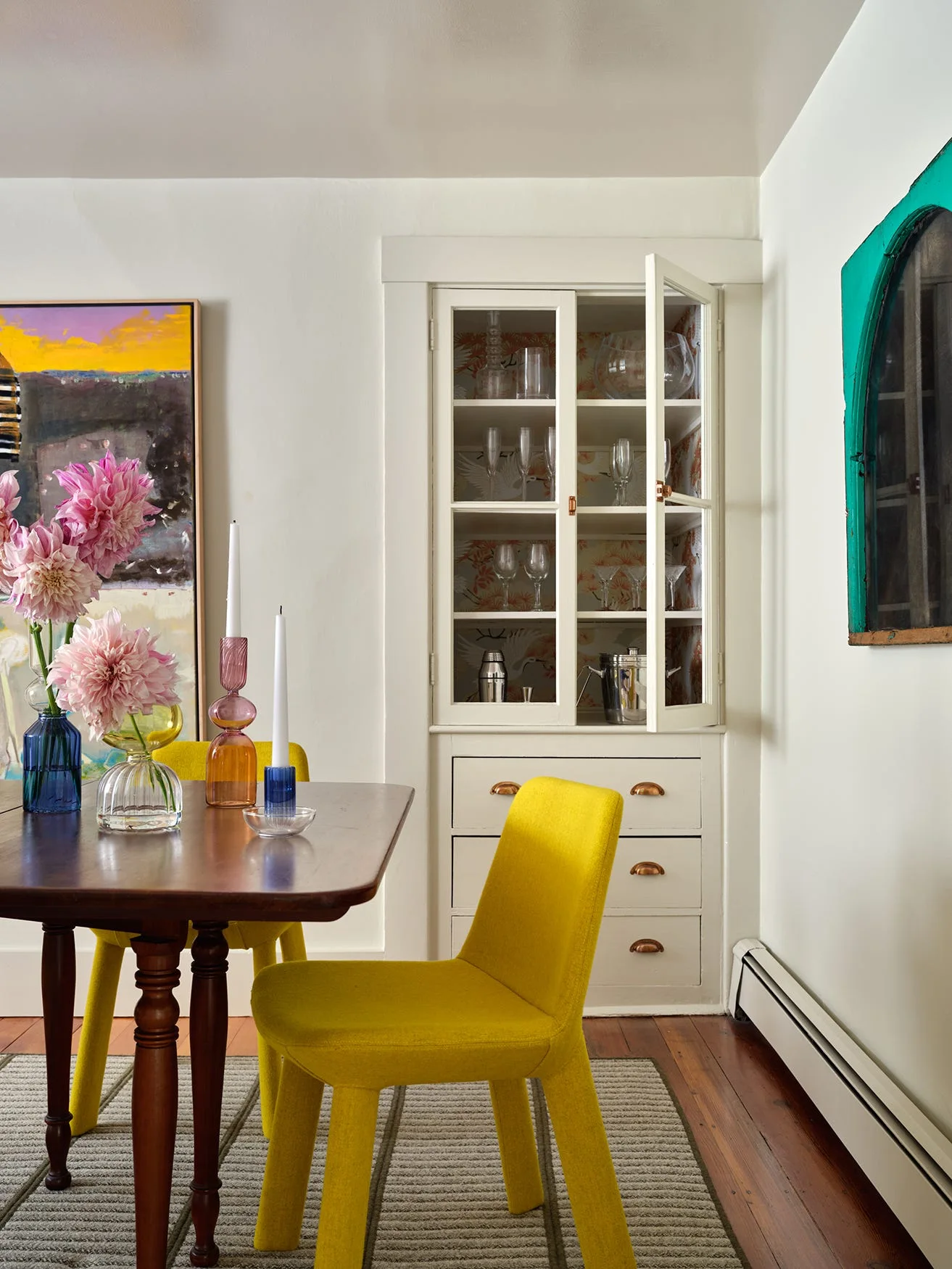 A dining room features yellow upholstered chairs around a dark wood table, a striped rug, and a white built-in cabinet backed with patterned wallpaper.