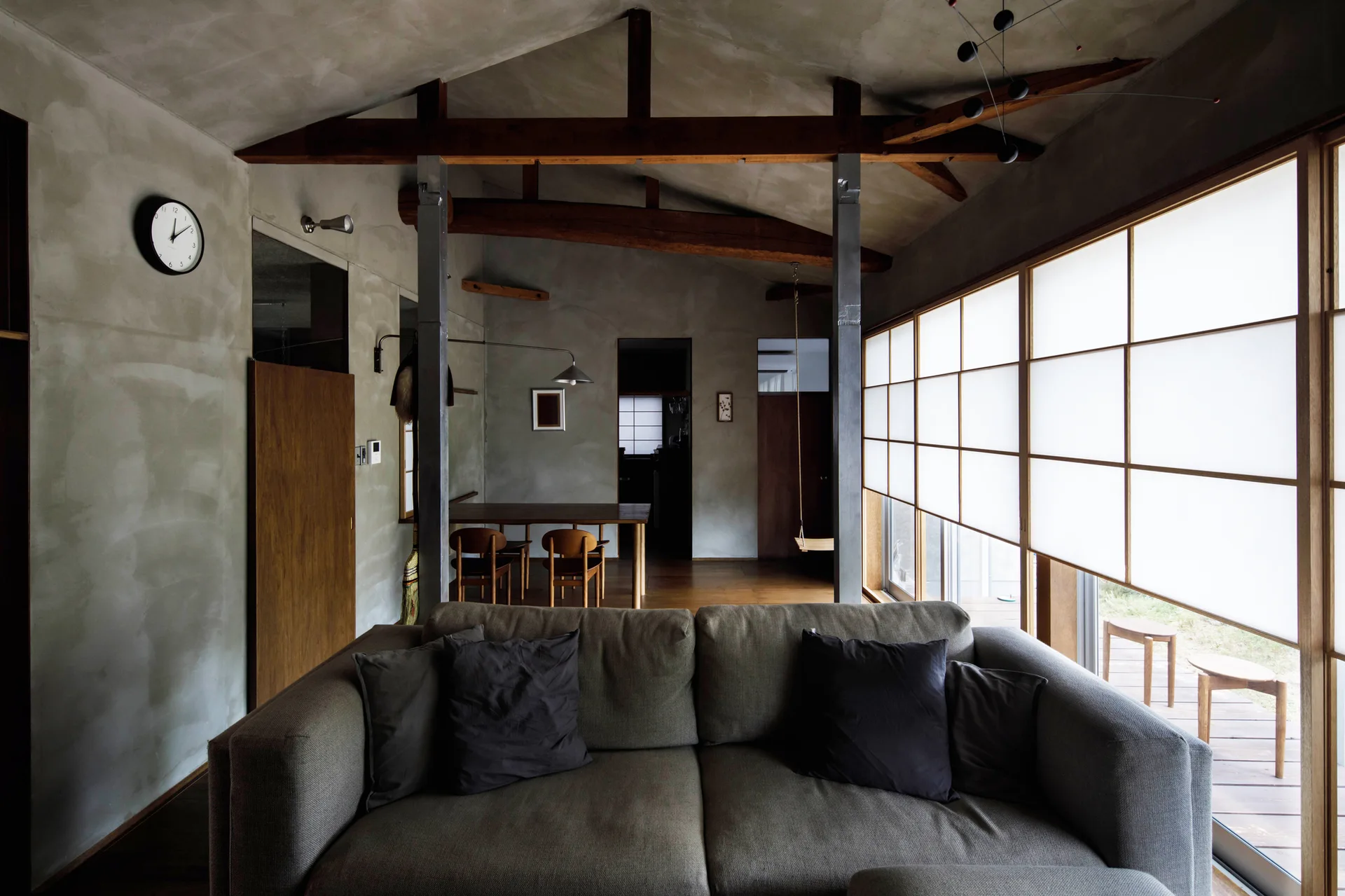 A modern living room with a grey sofa, exposed wooden ceiling beams, concrete walls, and large Japanese-style shoji screen sliding windows.