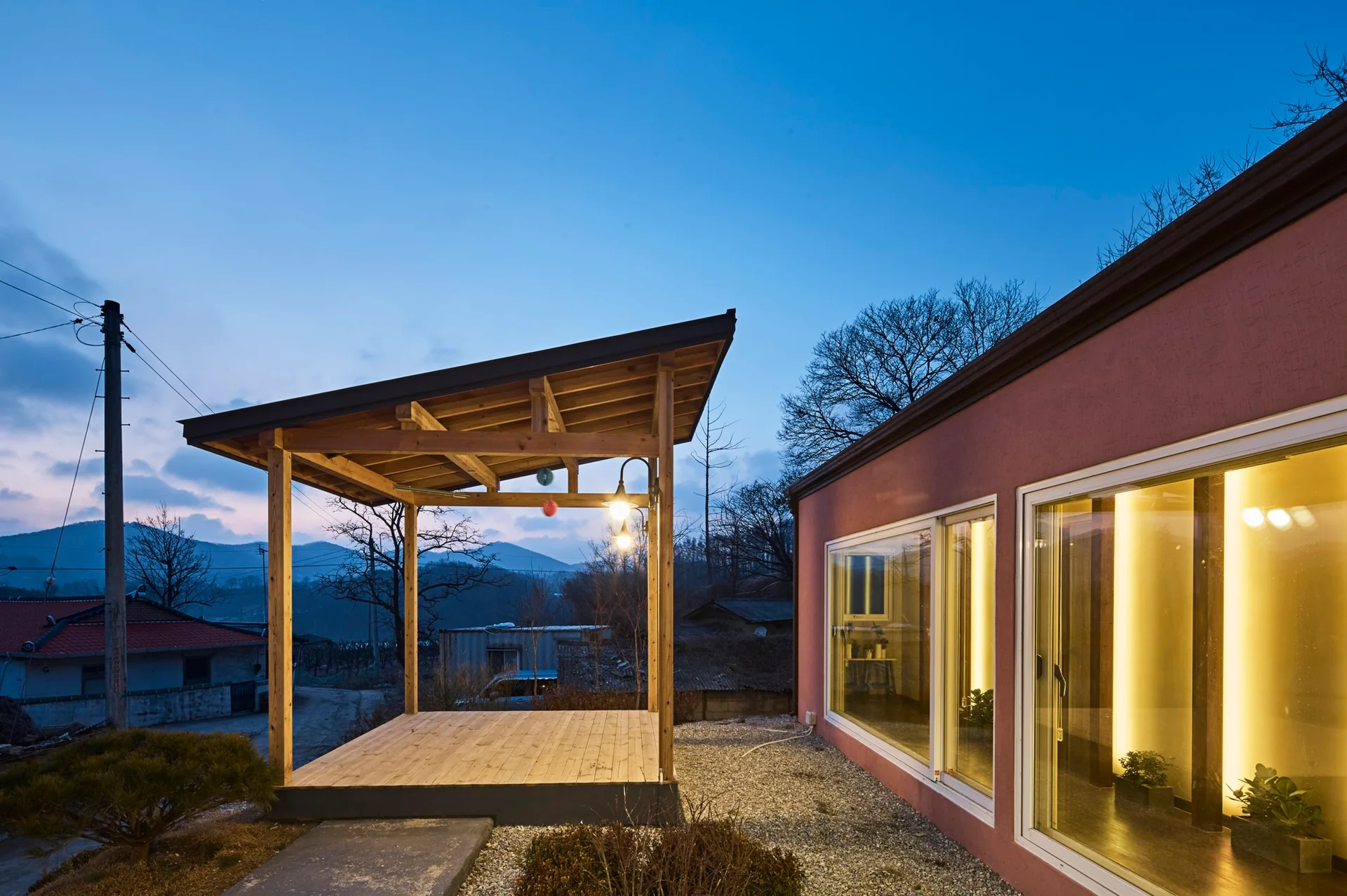 A detached covered wooden deck stands next to a terracotta stucco house with large illuminated windows under a blue twilight sky.