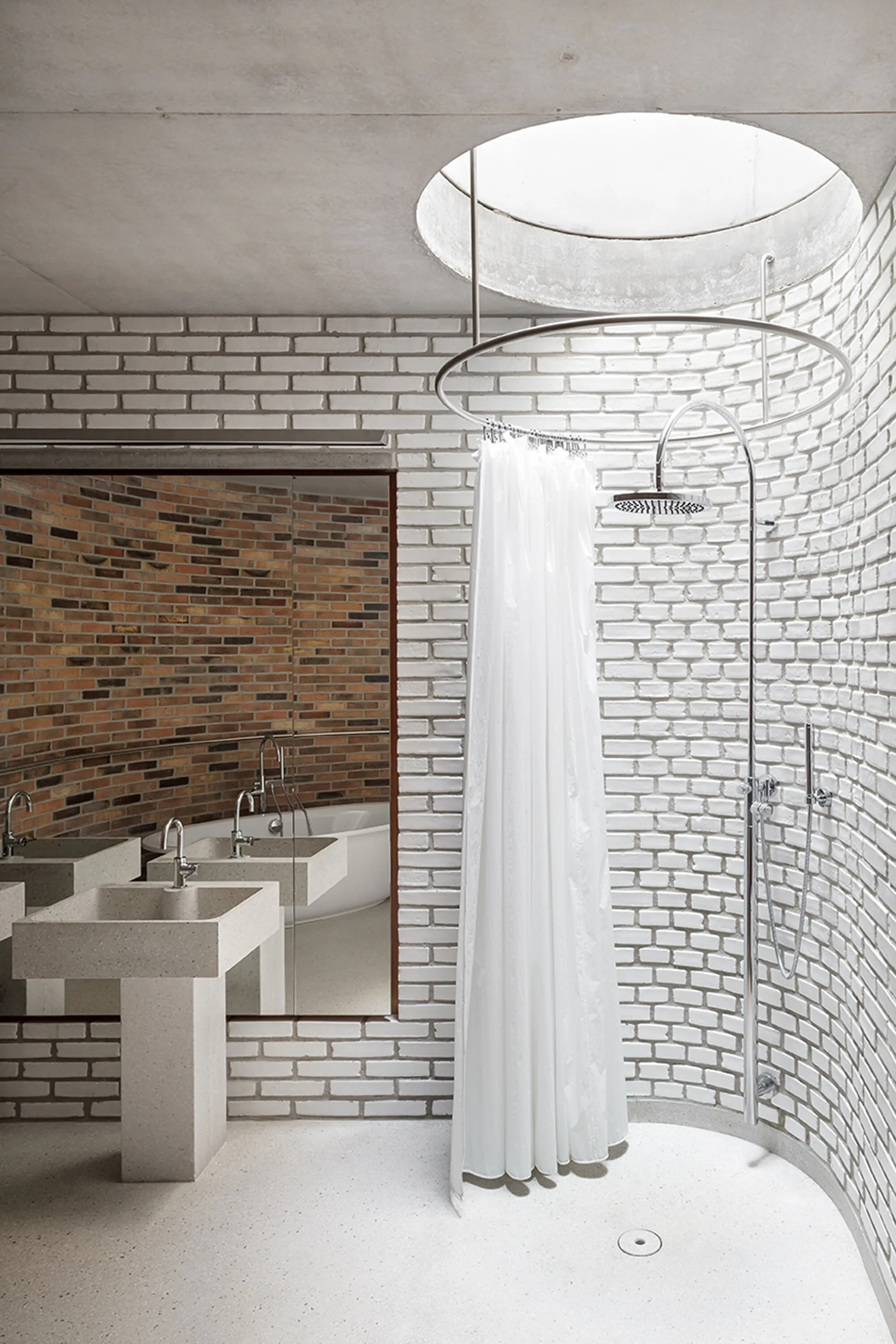 A minimalist bathroom featuring a curved white brick shower enclosure beneath a circular skylight next to a concrete pedestal sink.