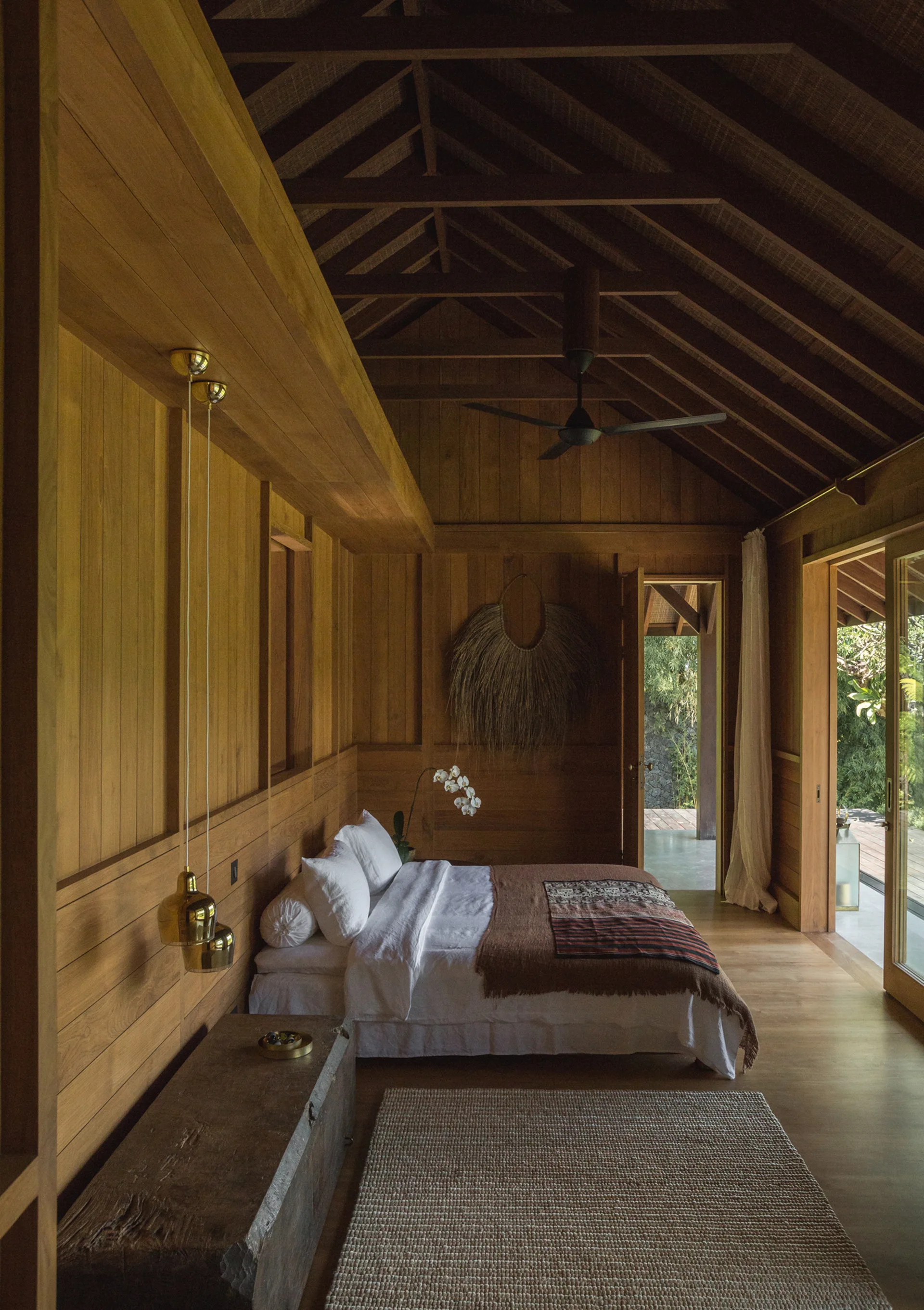 A bedroom features warm wood-paneled walls and a vaulted ceiling, a bed with white linens, gold pendant lights, and a woven rug.