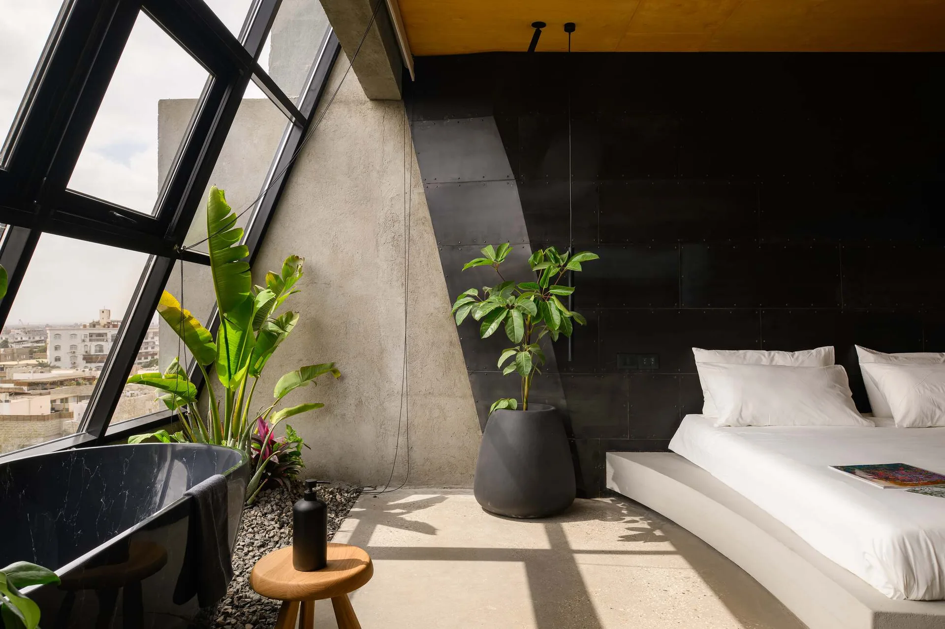 An industrial bedroom with a low concrete platform bed, a black metal accent wall, and a freestanding bathtub set on pebbles near a slanted window.