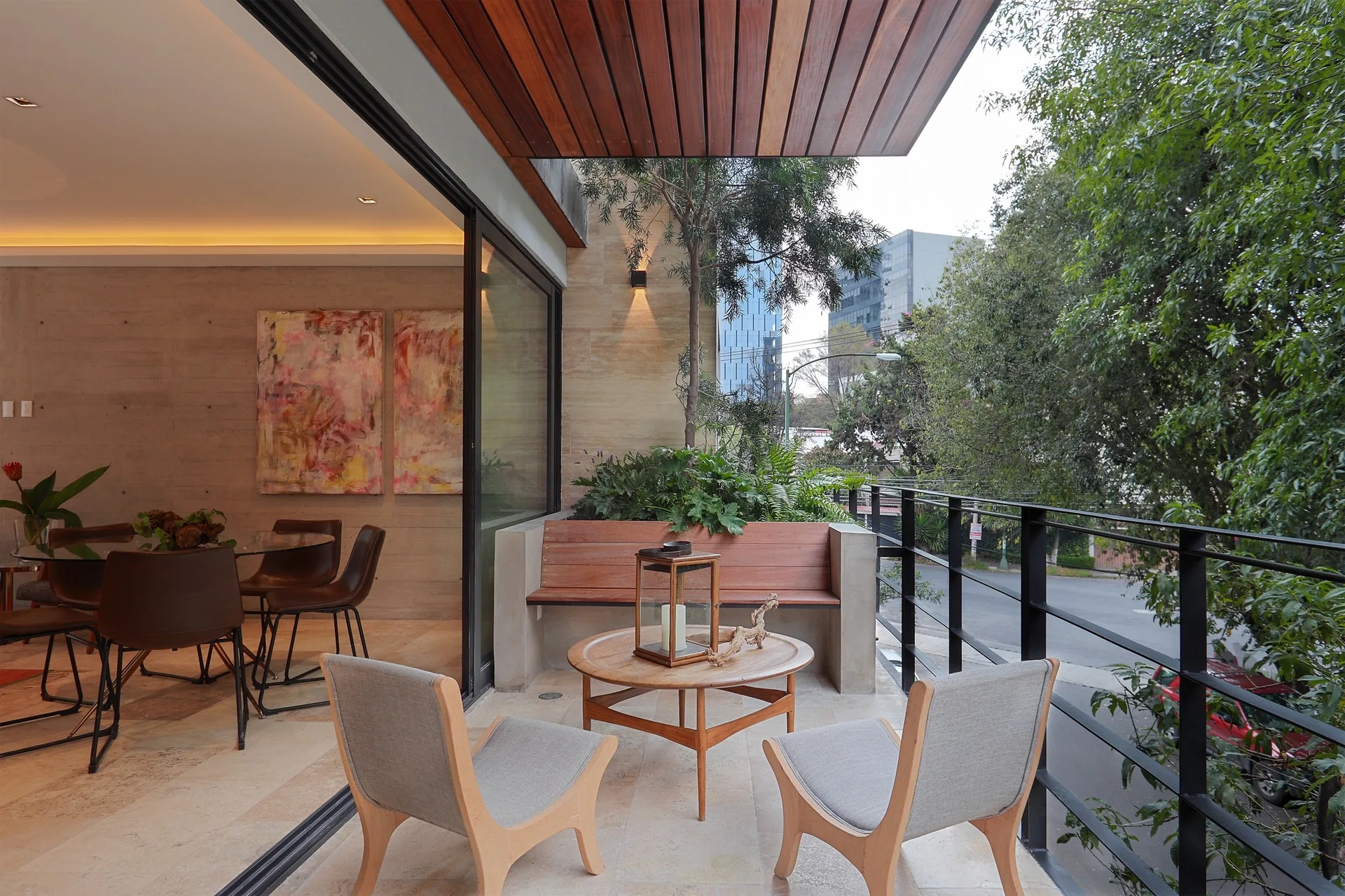 A modern apartment balcony features two grey lounge chairs, a wooden coffee table, a built-in bench, and sliding glass doors leading to a dining area.