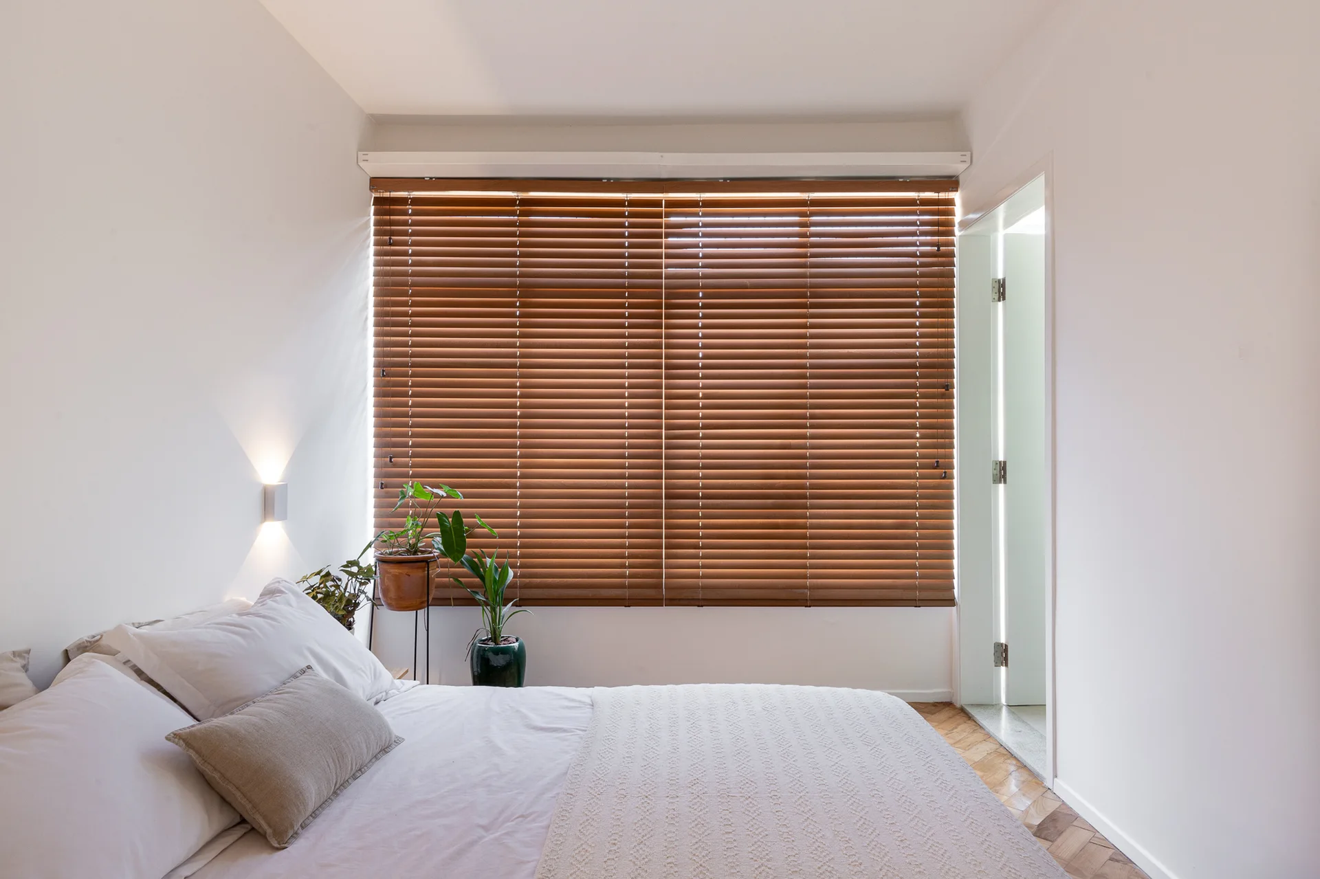 A minimalist bedroom features a bed with white linens, large brown wood blinds over the window, herringbone floors, and potted green plants in the corner.