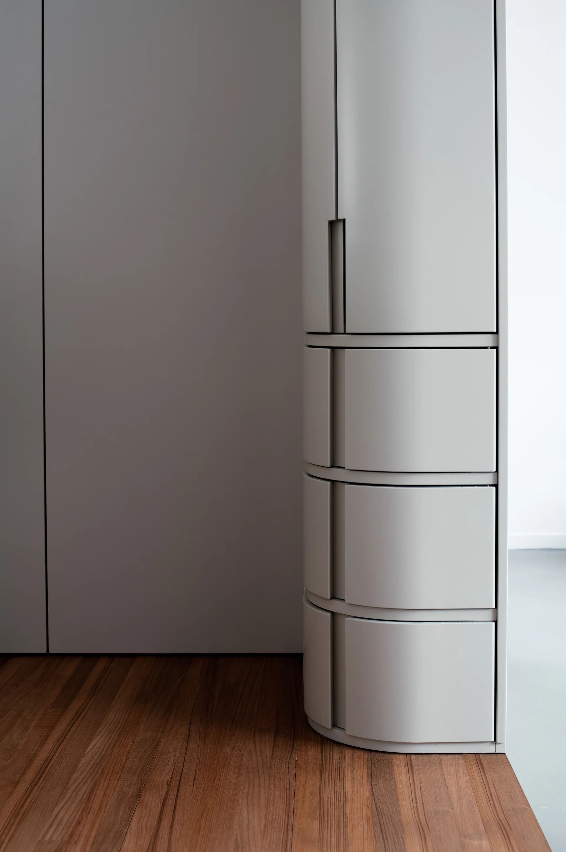 A minimalist light grey cabinet with a curved front and stacked drawers stands on a warm wood plank floor.