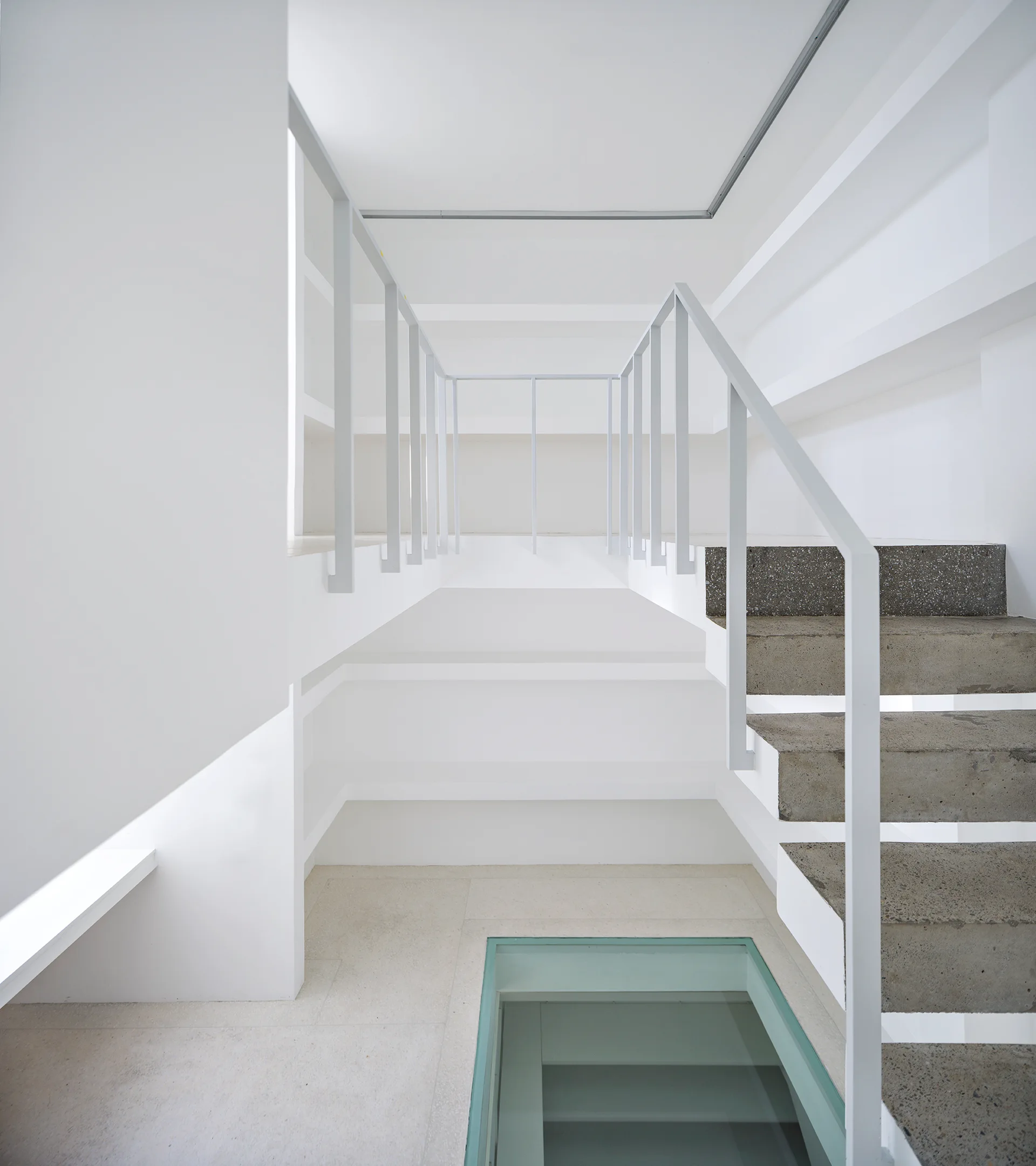 A minimalist white stairwell featuring raw concrete steps, a slender metal railing, and a rectangular glass floor insert.