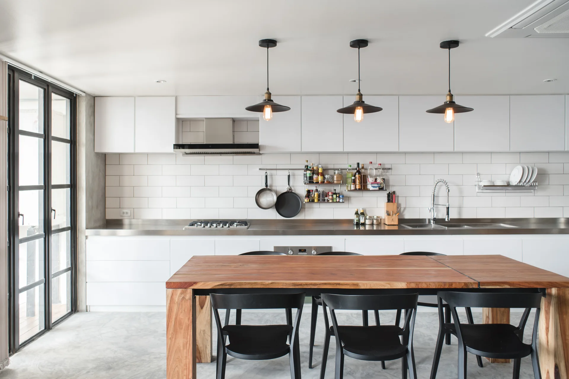 A minimalist kitchen featuring white cabinets, a stainless steel countertop with a subway tile backsplash, and a warm wooden dining table with black chairs.