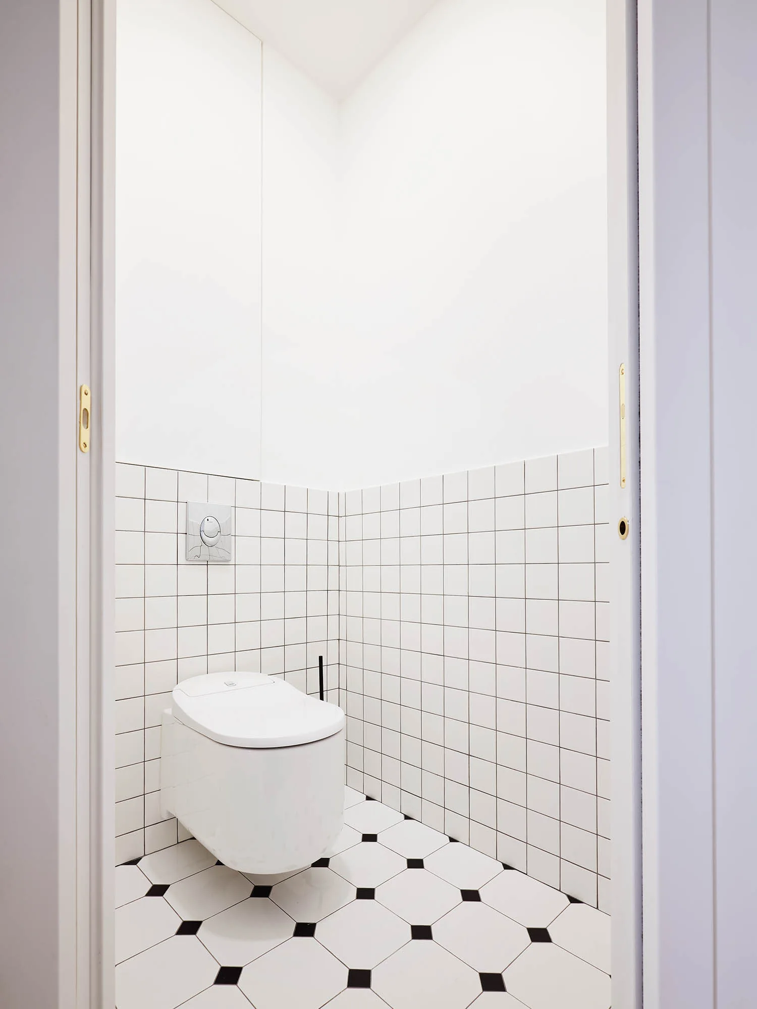 A minimalist powder room with a wall-mounted toilet, white square grid wall tiles, and a black and white geometric tiled floor.