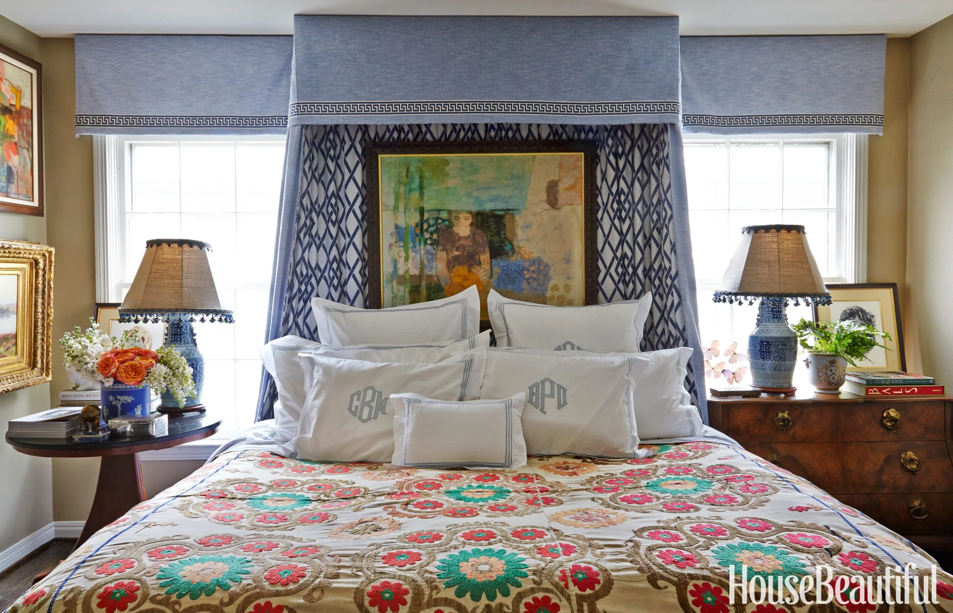A traditional bedroom features a bed with monogrammed pillows and a floral quilt centered between two windows under a blue fabric pelmet.