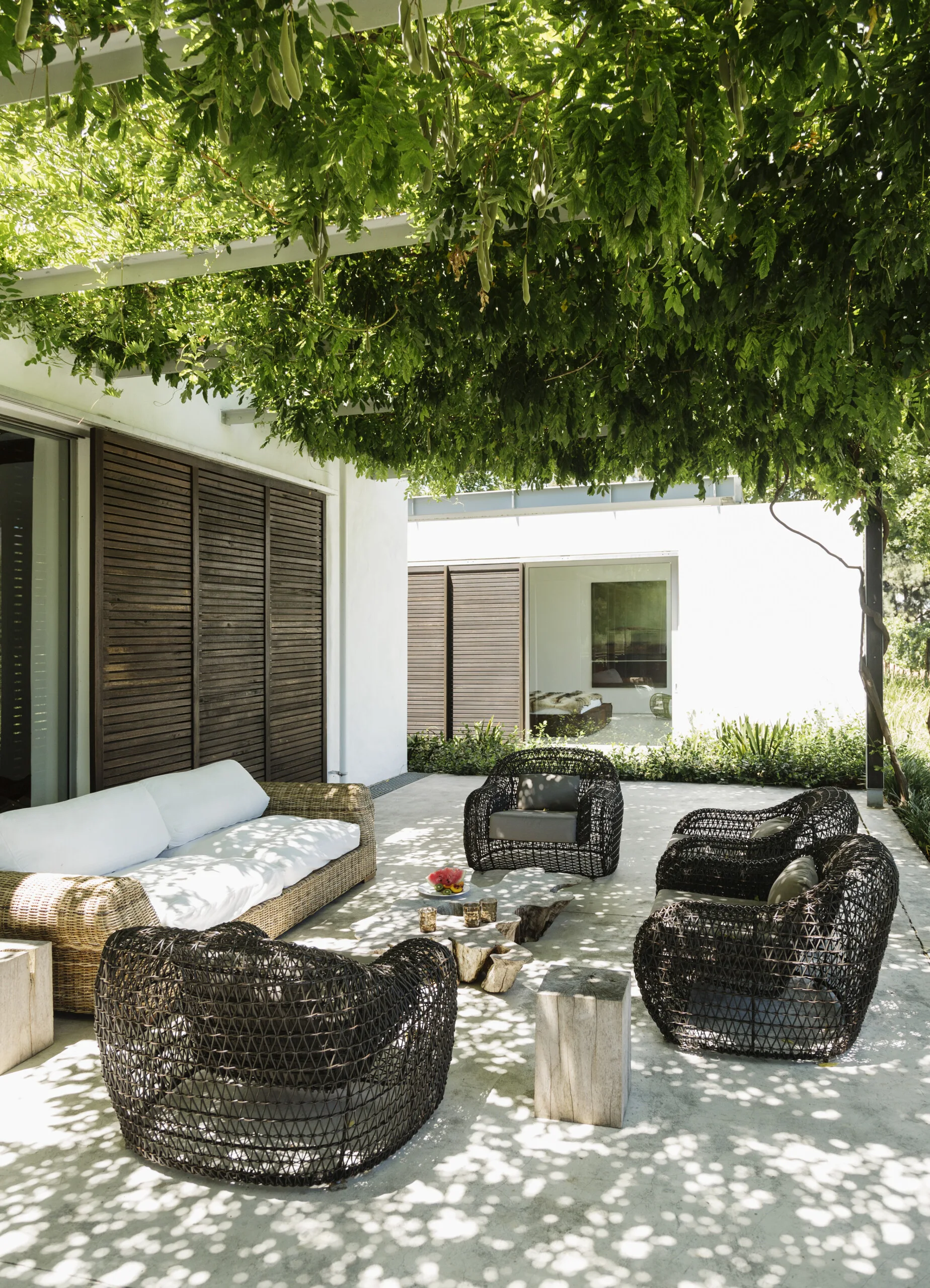 An outdoor paved patio features dark wicker seating with white cushions arranged beneath a lush green vine pergola.