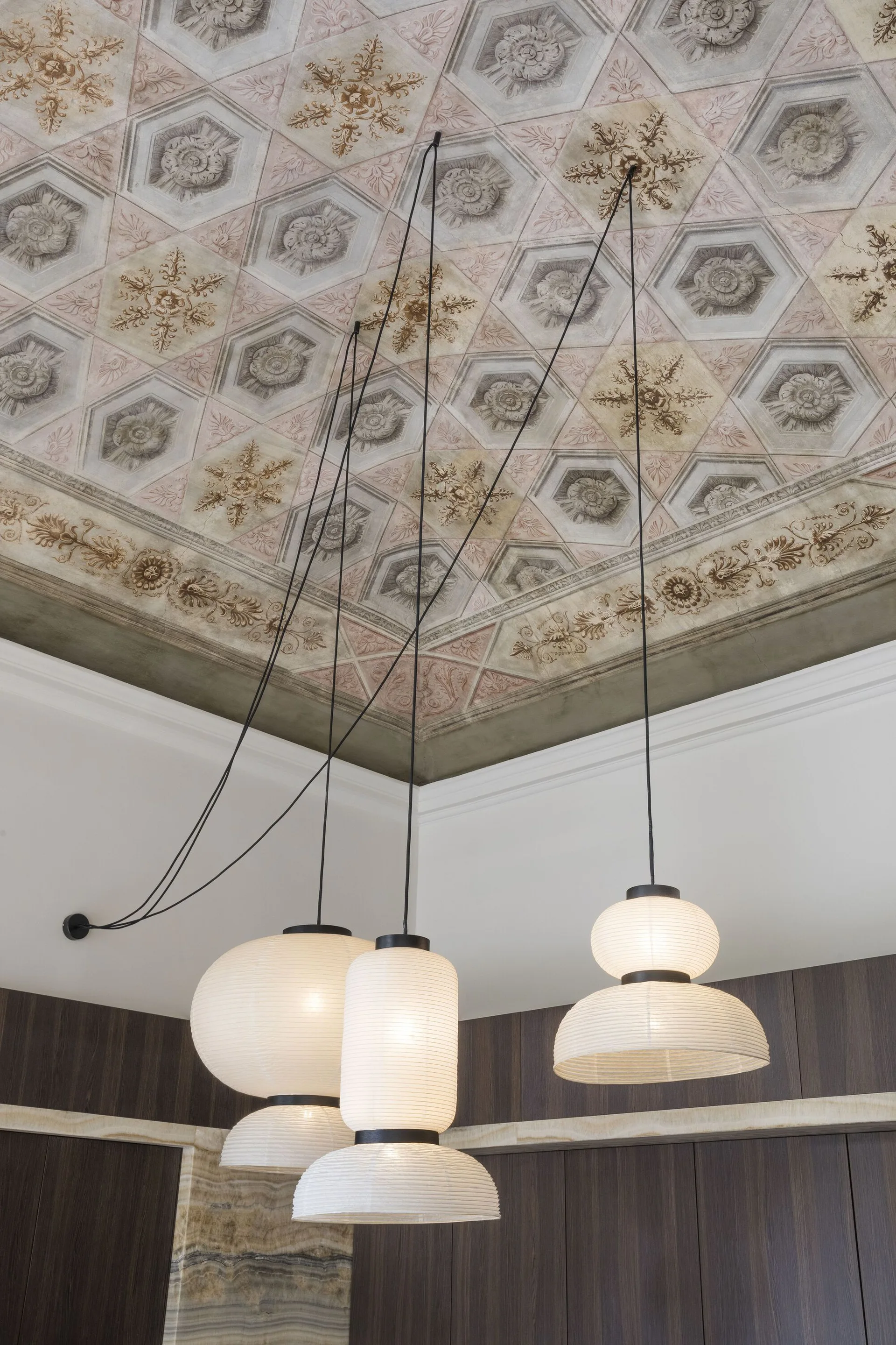 A cluster of glowing white paper lantern pendant lamps with black cords hangs beneath a highly detailed, hexagonal-patterned fresco ceiling.