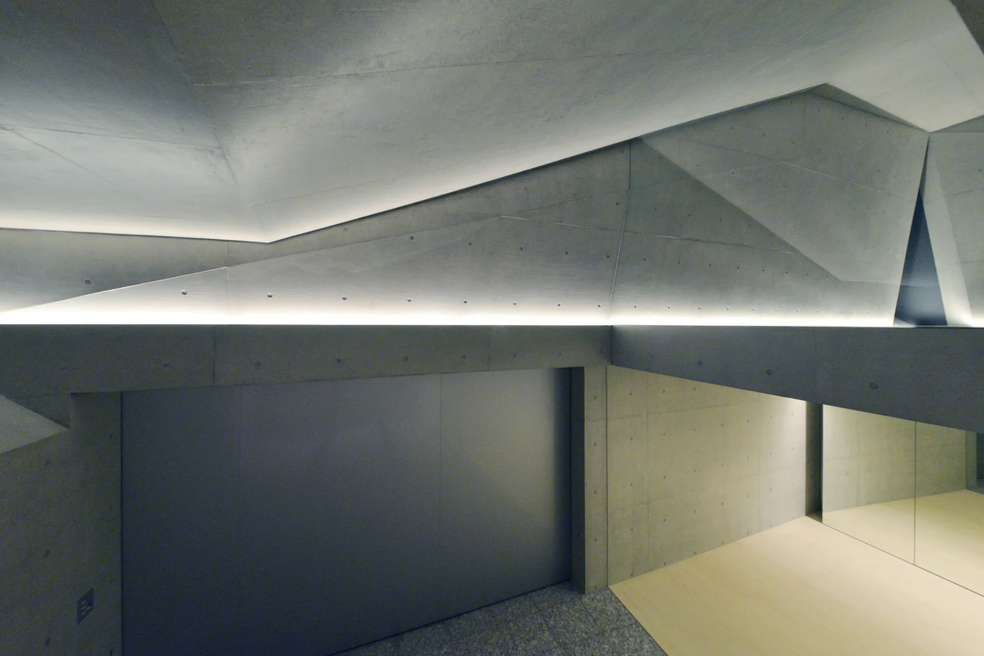 A minimalist interior with faceted concrete ceilings illuminated by integrated linear cove lighting above smooth grey concrete walls.