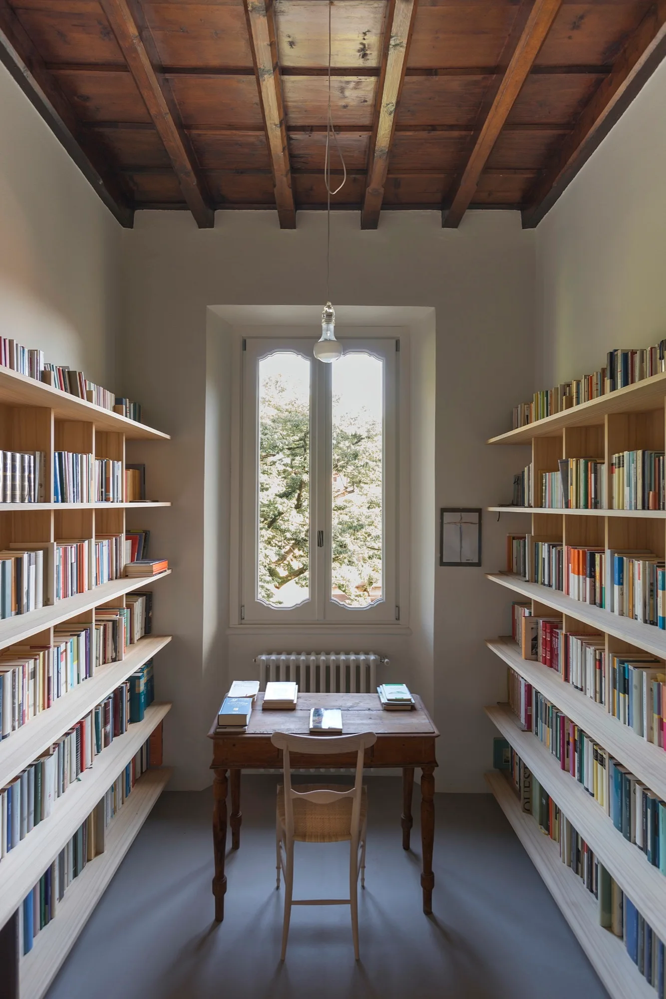 Symmetrical light wood bookshelves line a narrow home library with a wooden desk and chair centered before a window under an exposed beam ceiling.