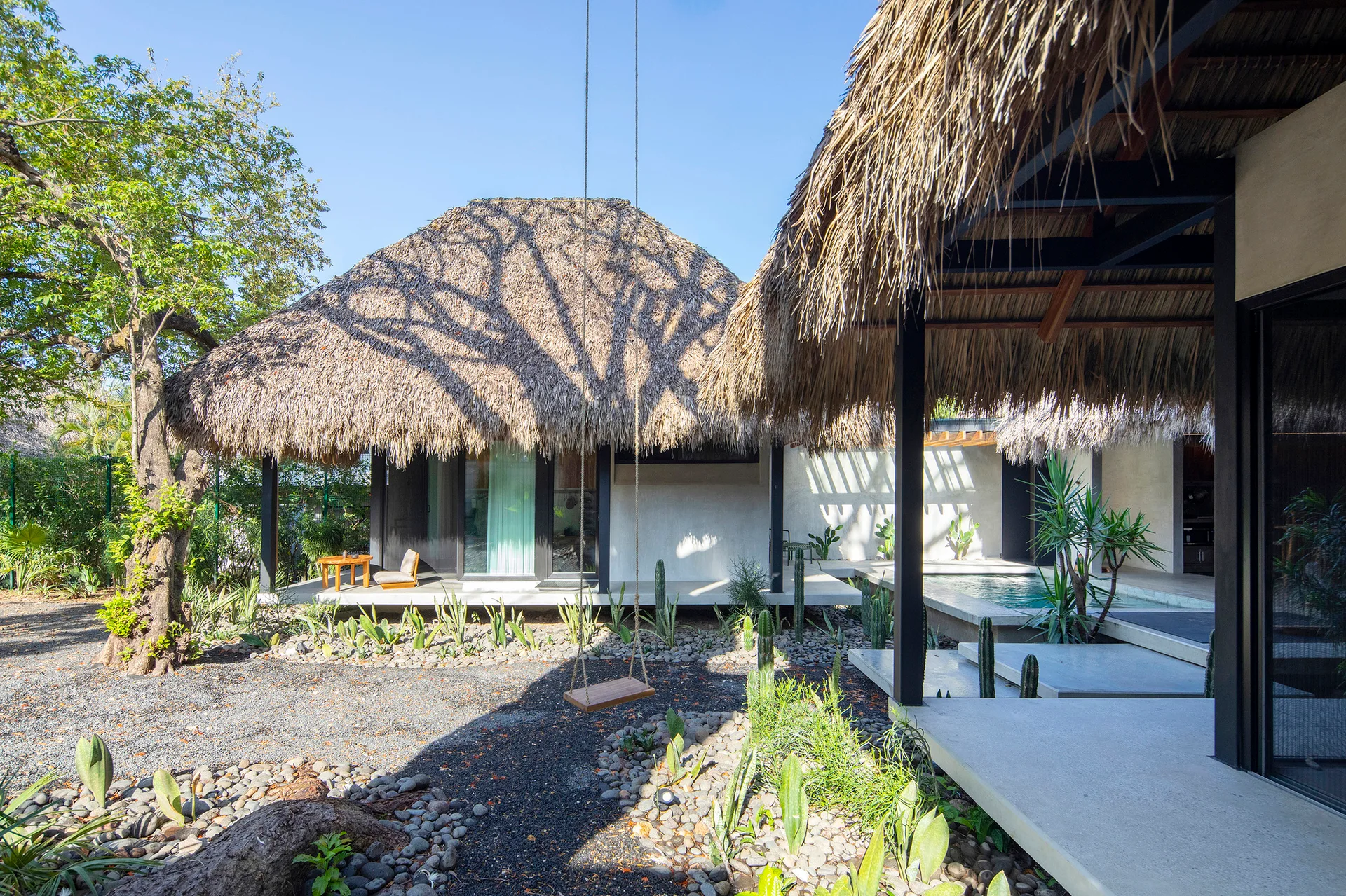 A modern tropical patio features a thatched roof structure, floating concrete platforms, a swimming pool, and a gravel garden with cacti and a rope swing.