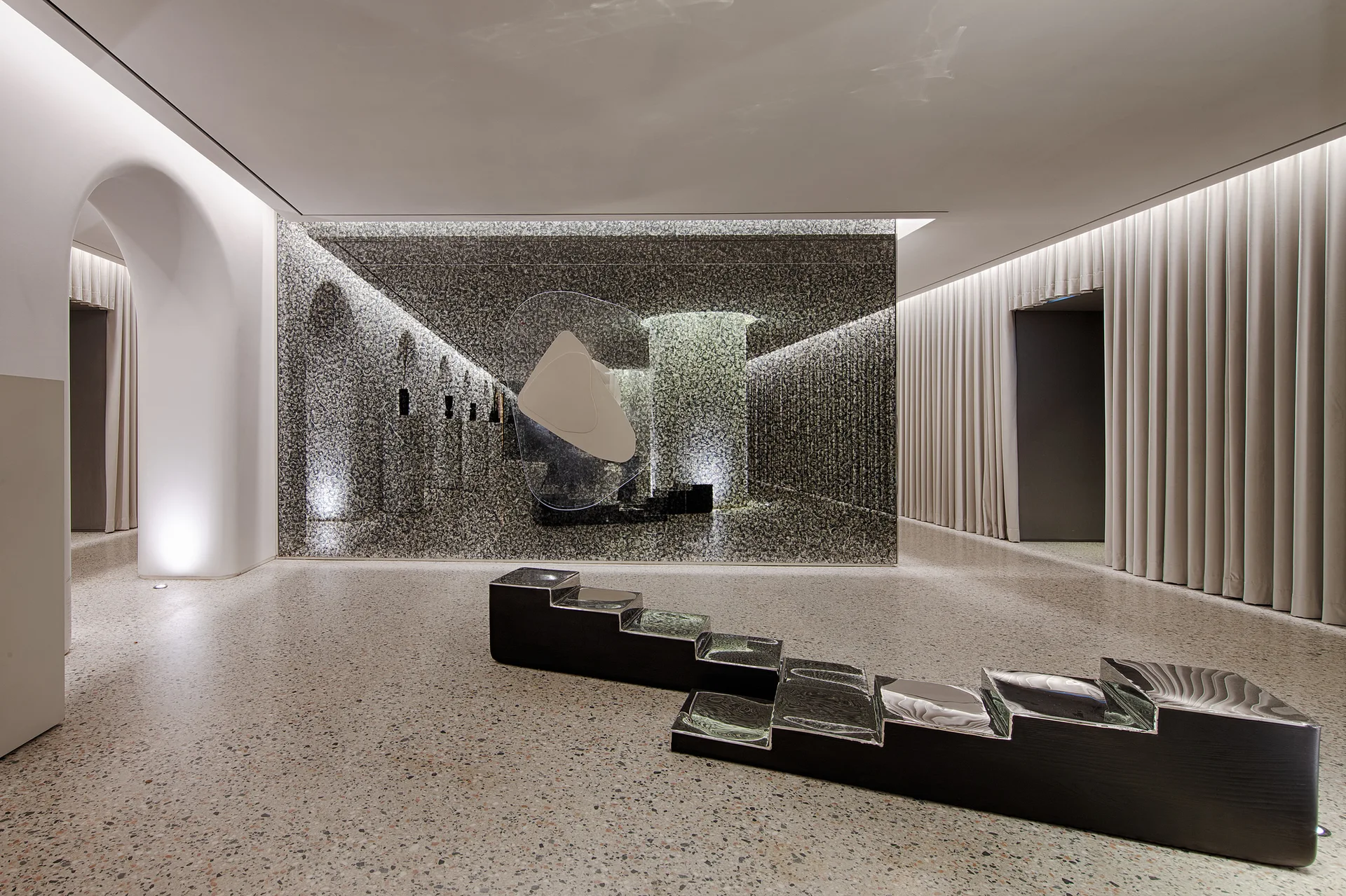 A minimalist interior lobby features a black tiered sculptural bench on terrazzo flooring in front of a mottled grey stone feature wall.