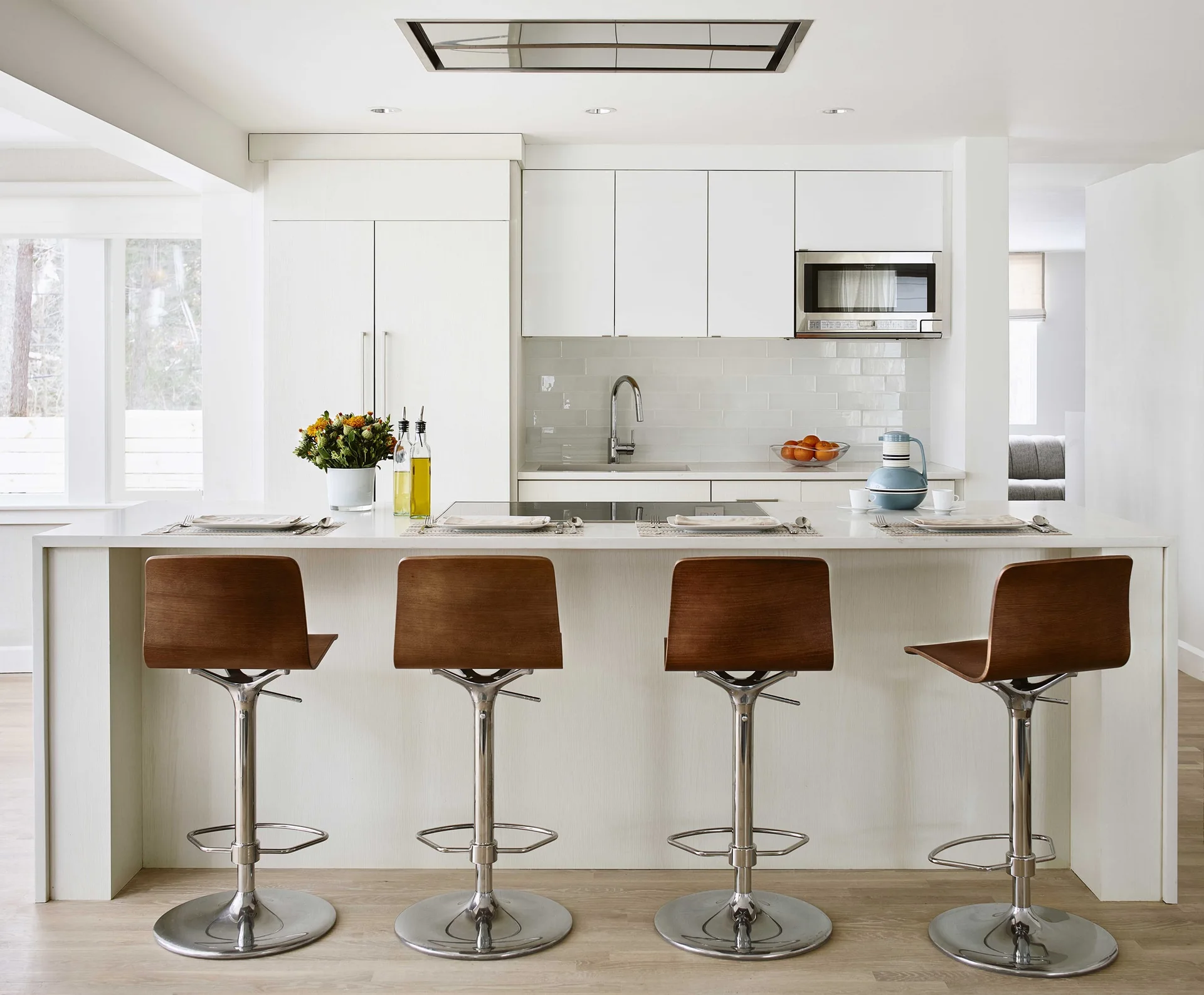 Four wood-toned bar stools line a long white kitchen island beneath a subway tile backsplash and glossy white upper cabinets.