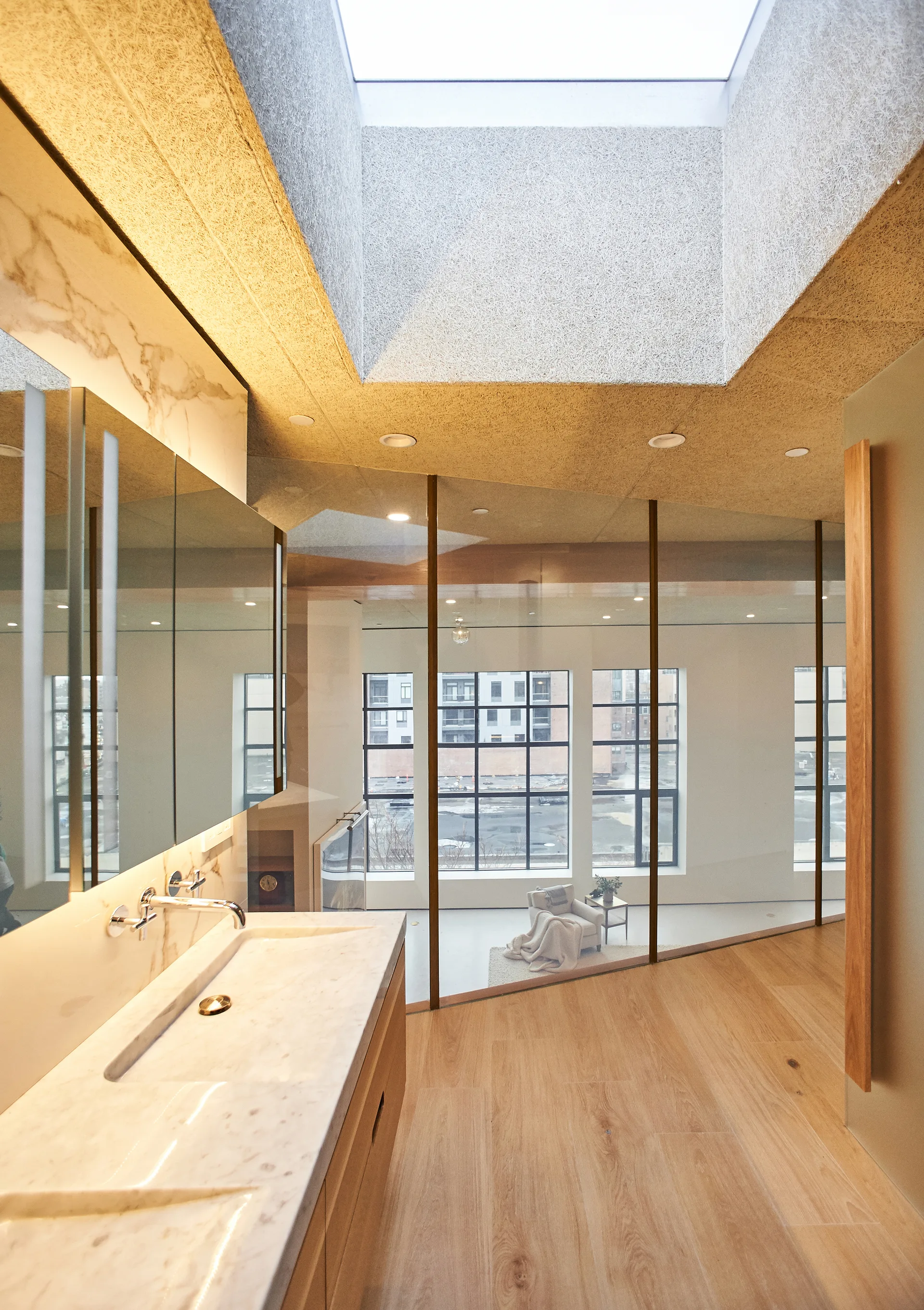 A modern bathroom features a veined marble vanity and a glass partition wall overlooking a living space lit by a large skylight.