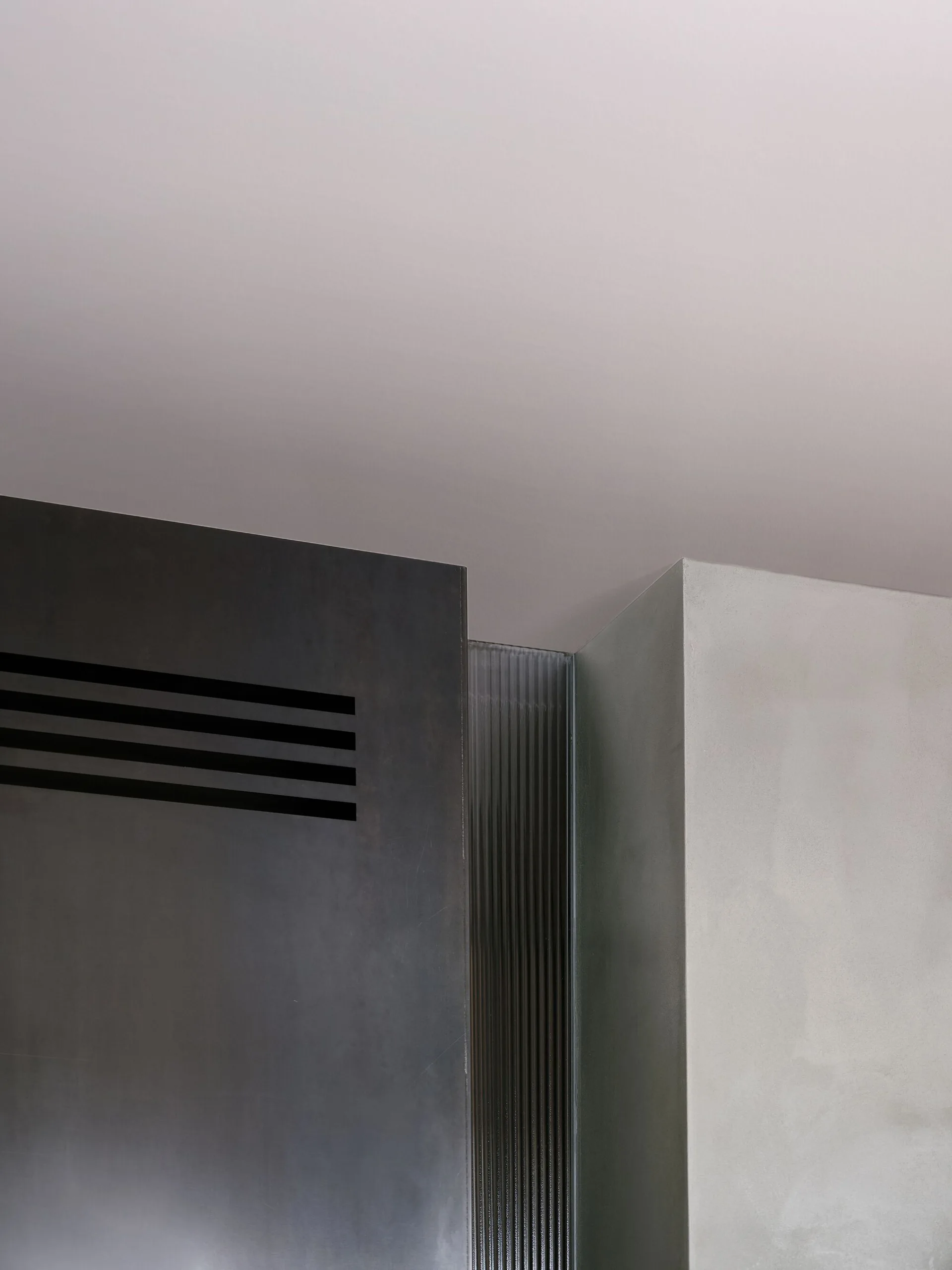 A close-up of a minimalist wall junction featuring a dark slatted metal panel, a vertical strip of fluted glass, and a light gray plaster wall.