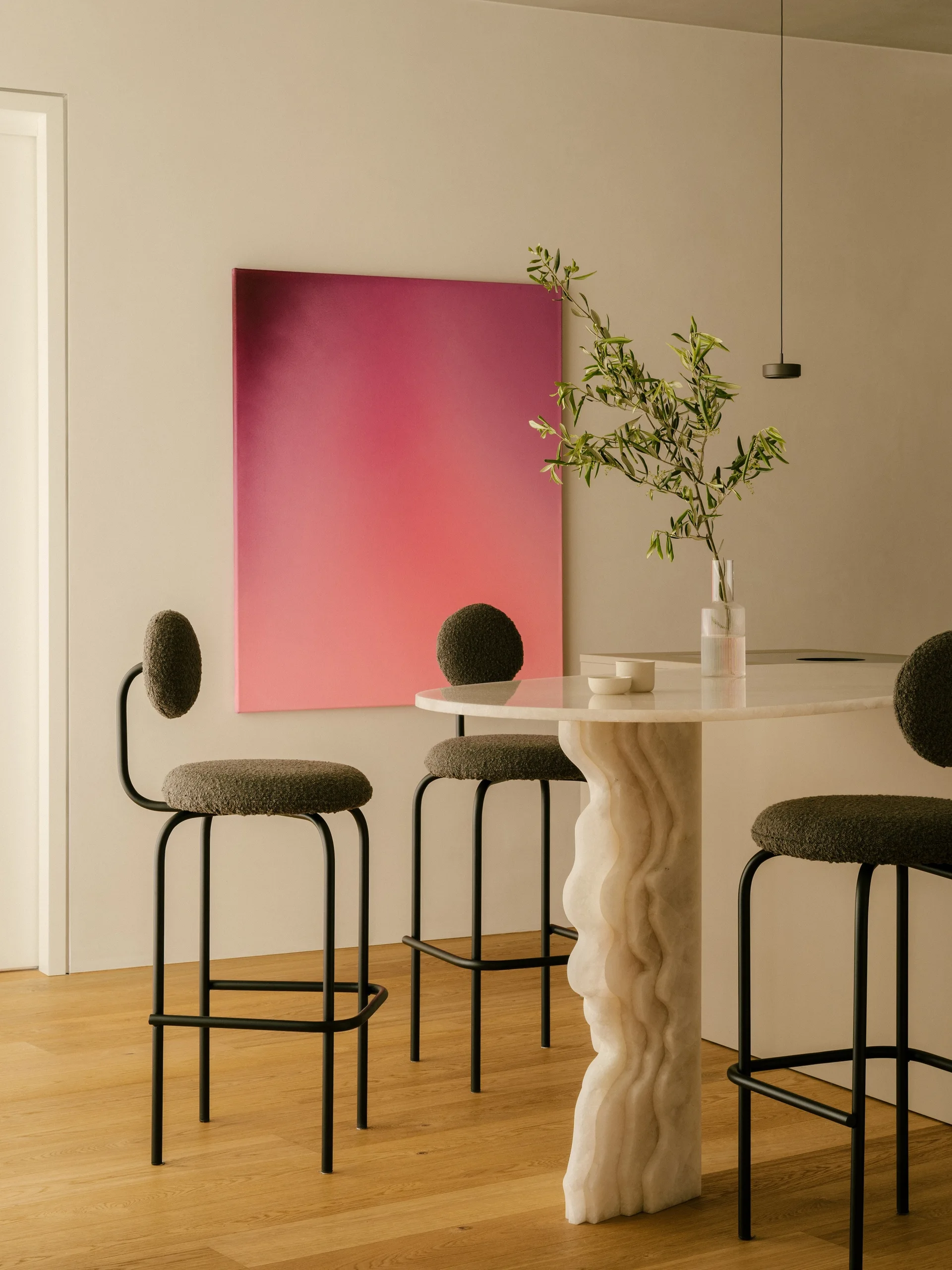 Three dark boucle bar stools surround a sculptural stone pedestal table set against a beige wall displaying a large pink gradient painting.