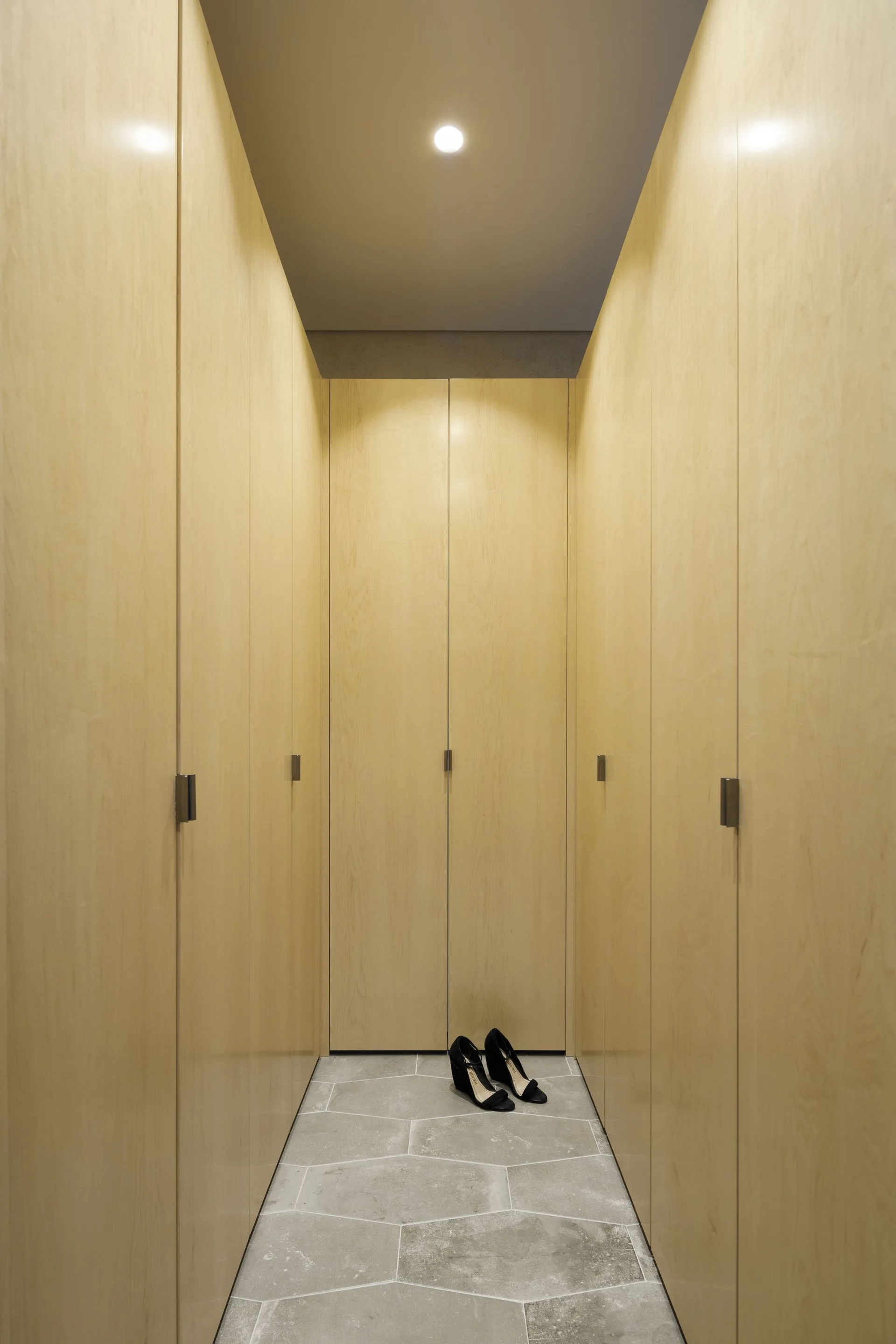 A narrow walk-in closet featuring full-height light wood wardrobe doors on both sides and a geometric grey stone tiled floor.