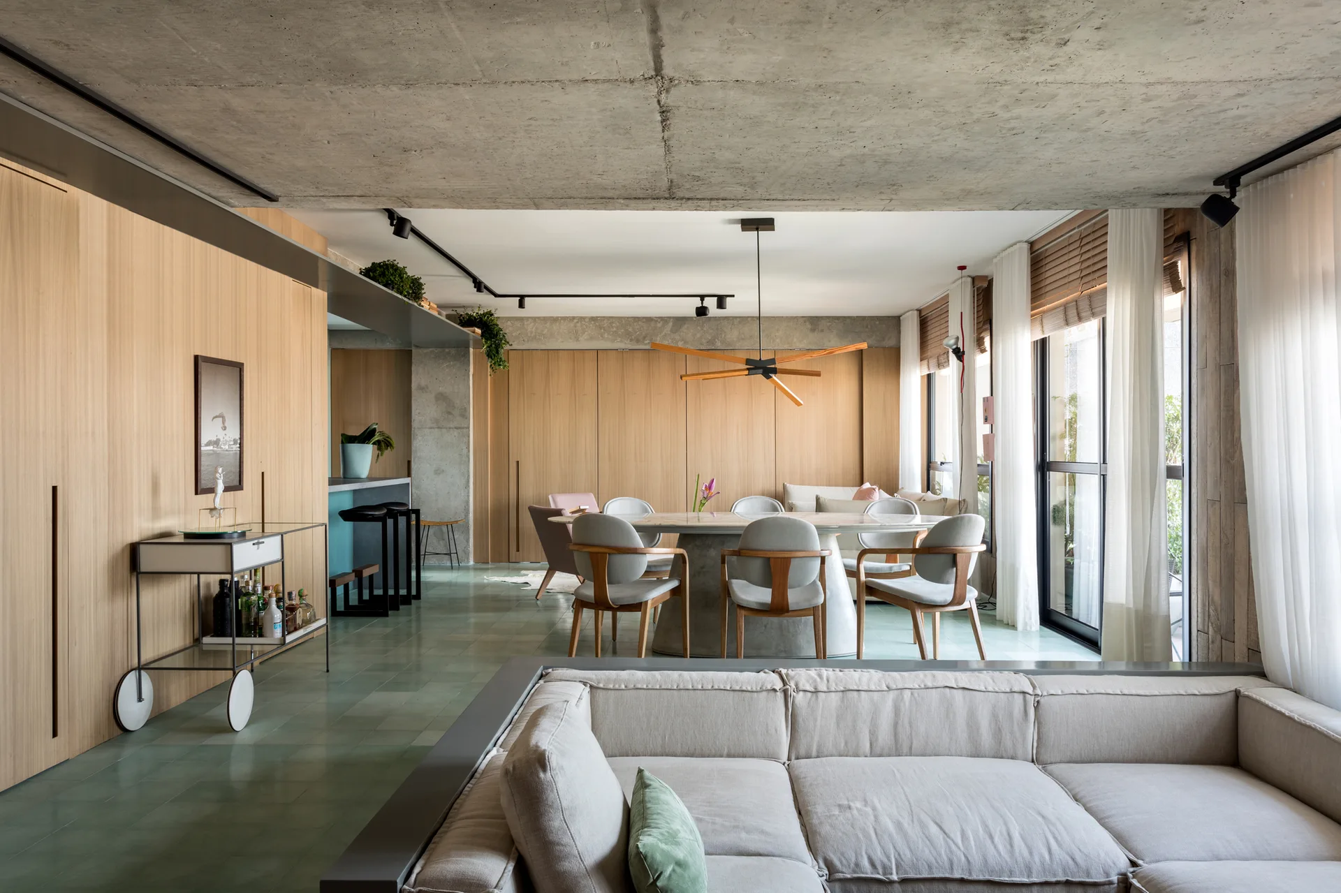 An open-plan apartment features an oval dining table, a light grey sofa, floor-to-ceiling wood paneling, and sage green tile flooring.