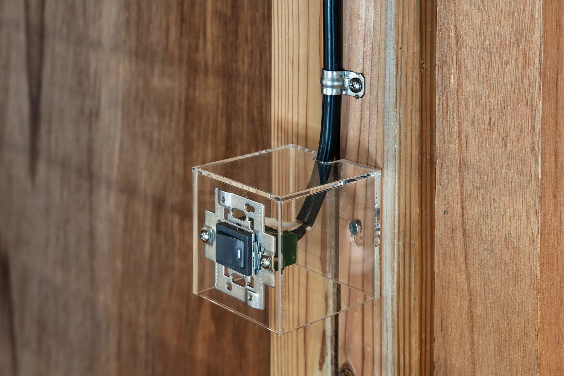 A black electrical rocker switch is enclosed in a clear acrylic box and surface-mounted on an exposed wood stud.