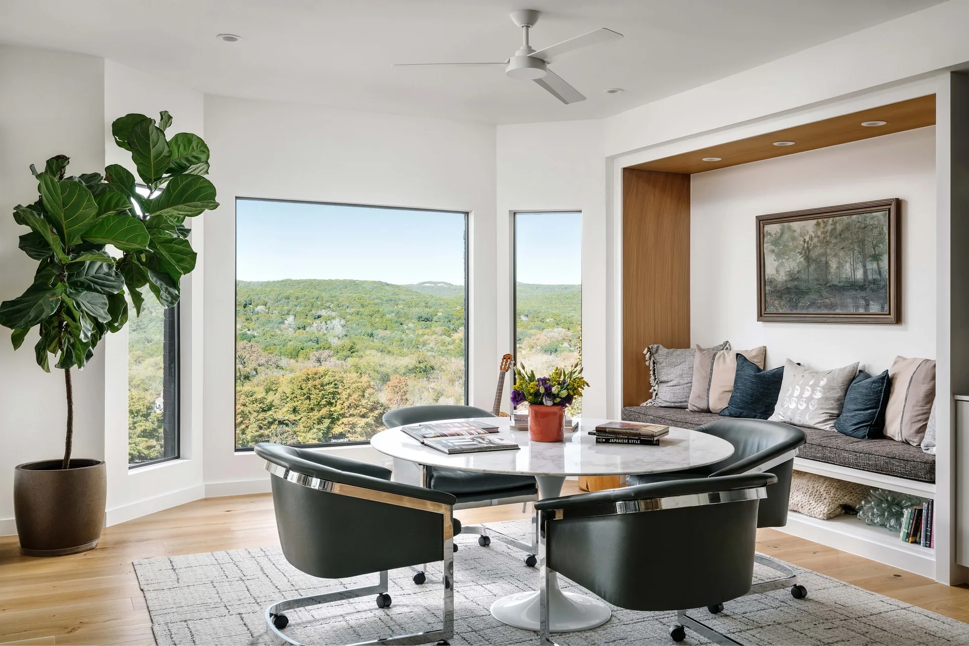 A modern dining room features a round marble table with dark grey swivel chairs next to a large window and a built-in recessed wooden bench.