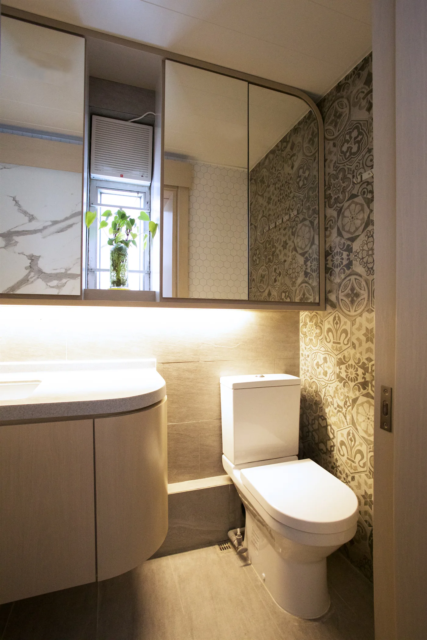 A compact modern bathroom featuring a curved wood vanity, a white toilet, patterned accent wall tiles, and mirrored cabinets with under-lighting.