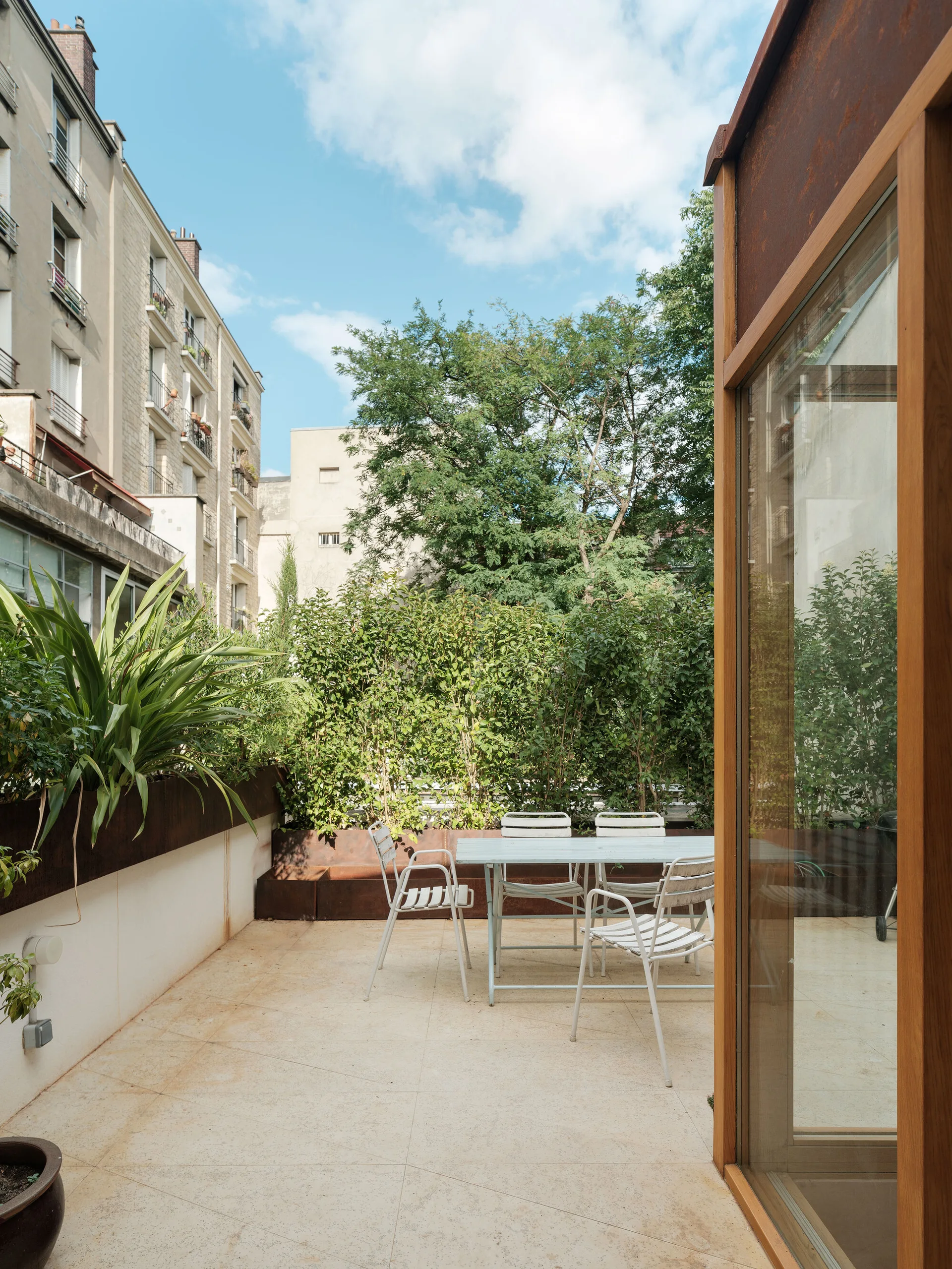 A metal dining table and chairs sit on a light beige tiled terrace bordered by lush green hedges under a blue sky.