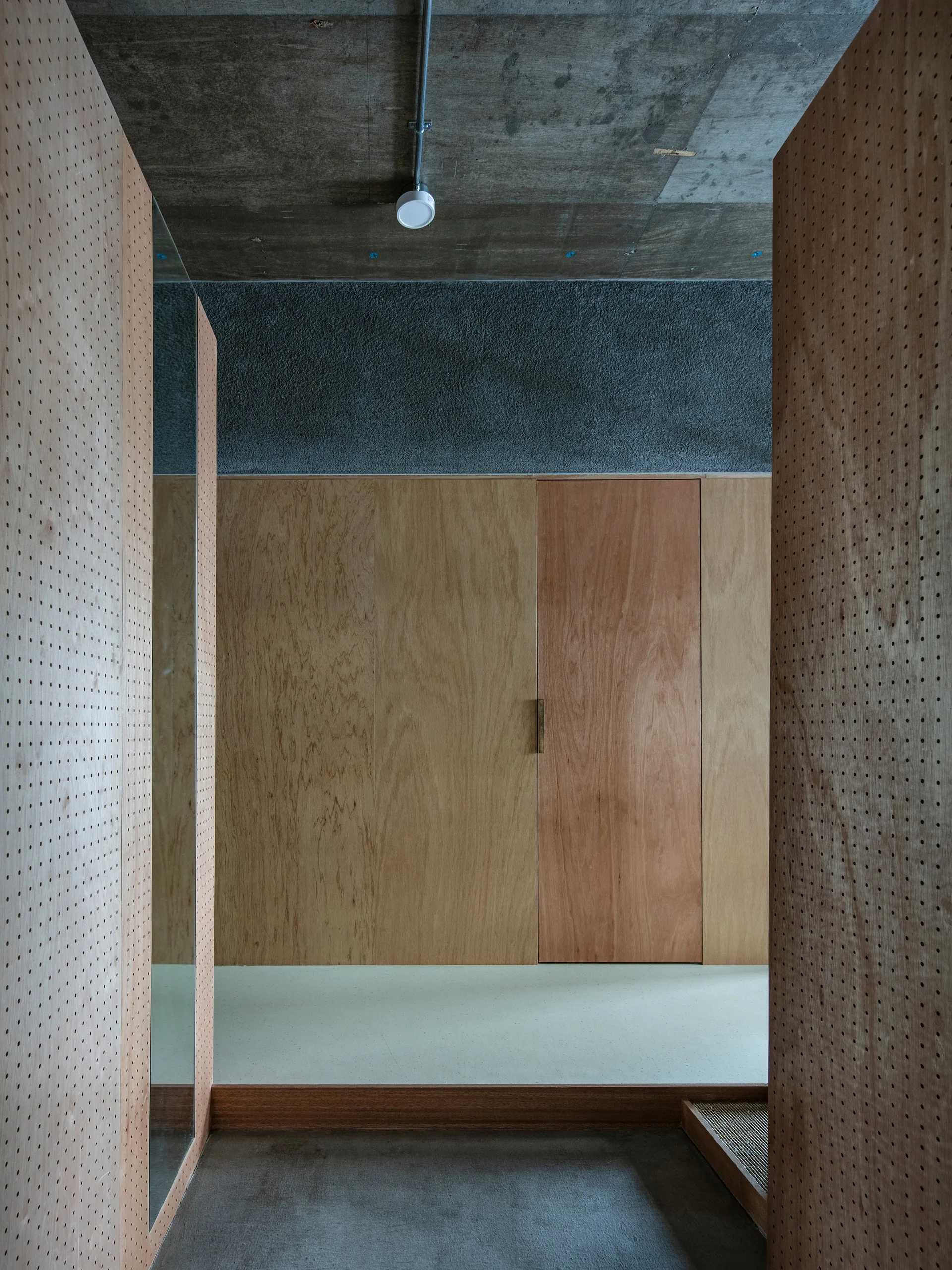 A minimalist hallway features walls made of perforated and smooth plywood panels beneath a raw gray concrete ceiling with a simple light fixture.