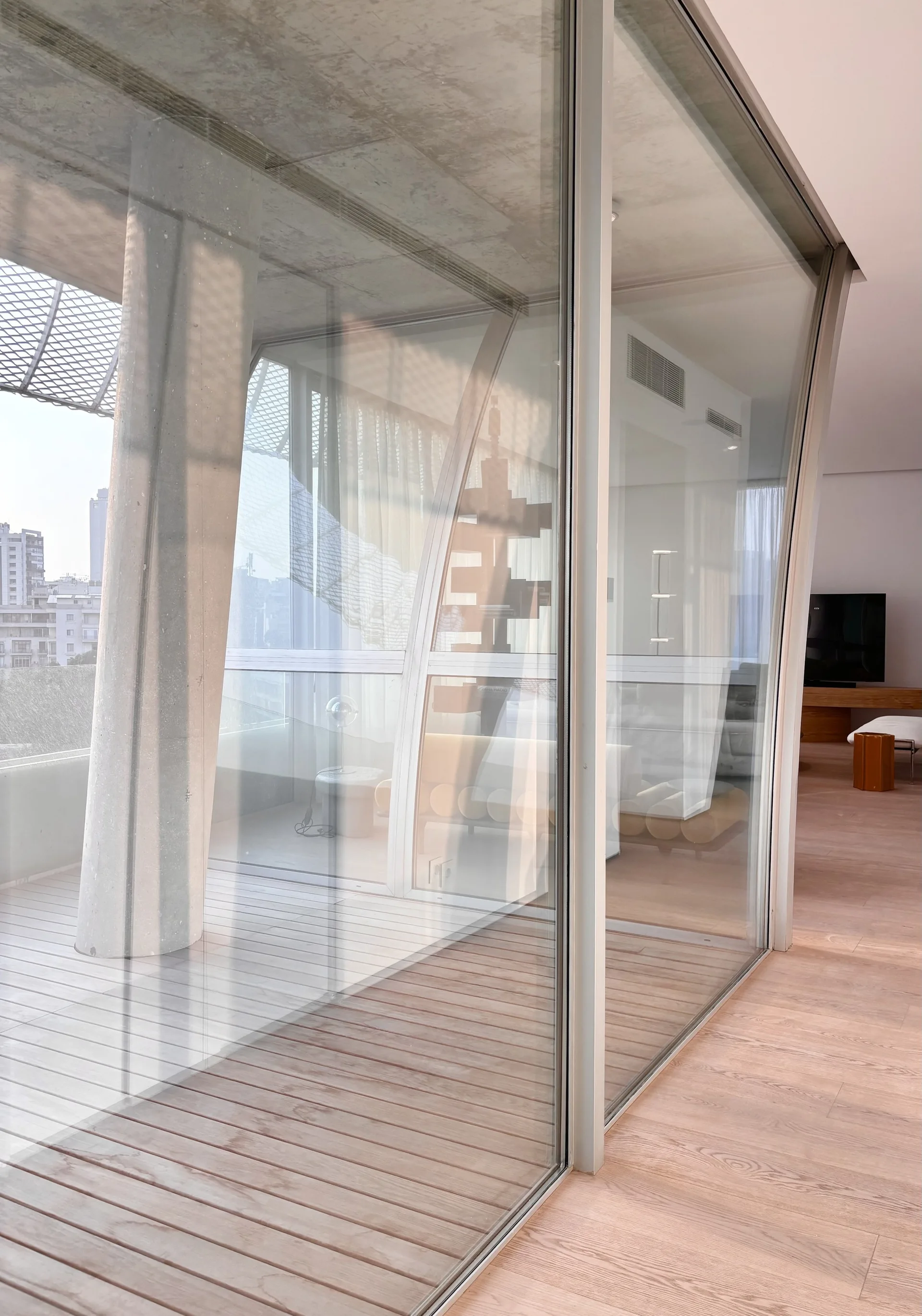 A minimalist interior space with a floor-to-ceiling glass wall separating a light wood living room from an outdoor balcony with a concrete column.