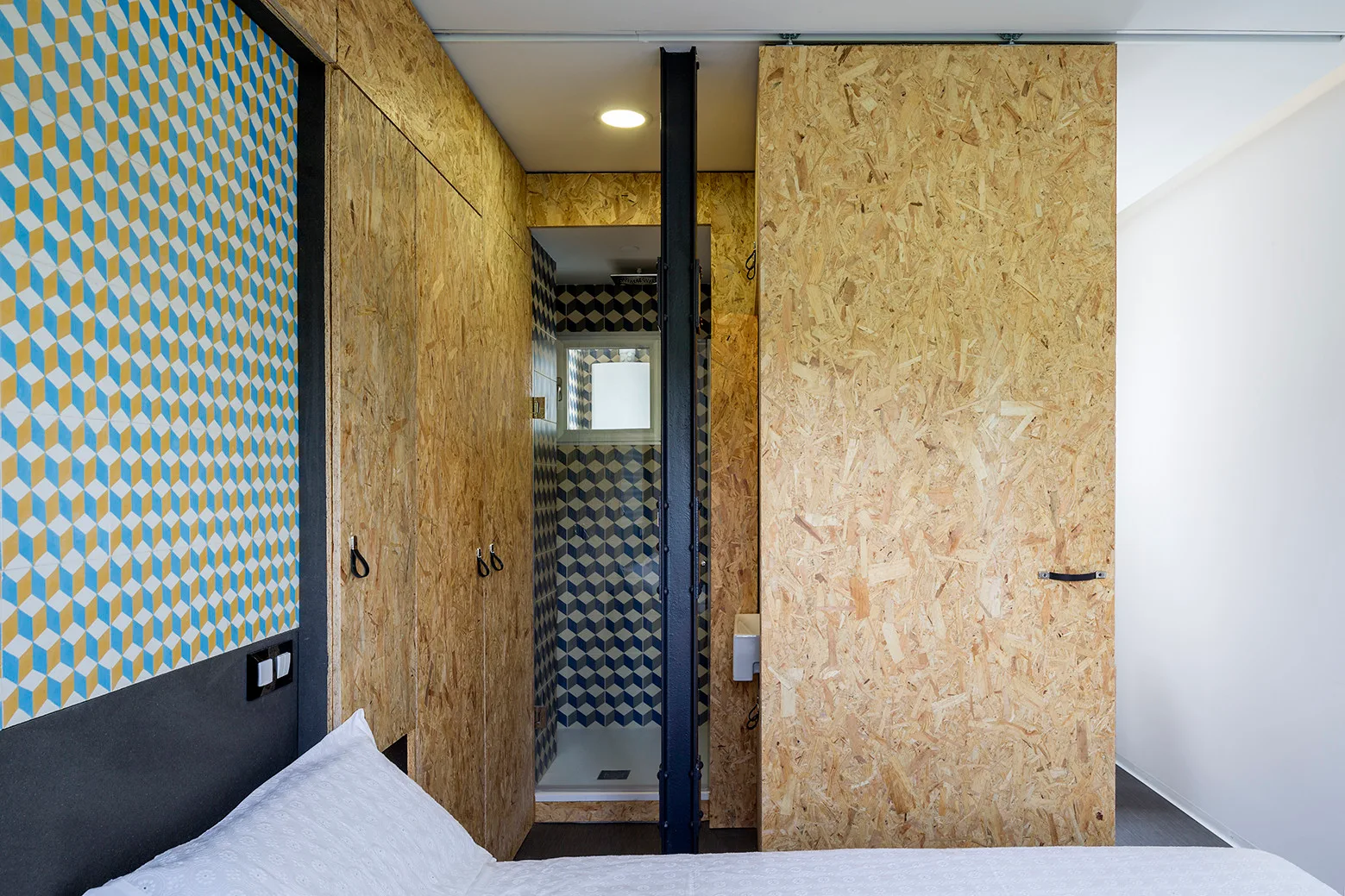 A modern bedroom features raw OSB wall panels, a dark structural metal column, and an adjacent bathroom with blue and yellow geometric tile.