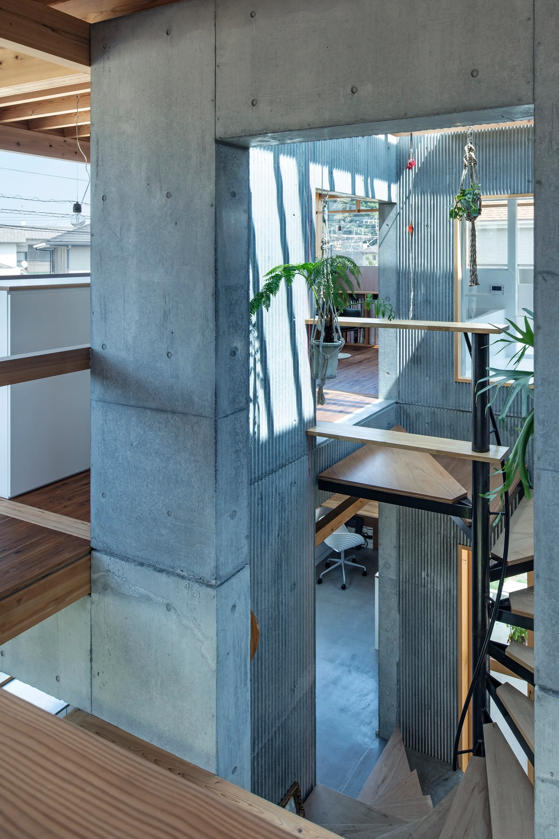 A multi-level interior showing a wooden staircase surrounded by raw concrete pillars, corrugated walls, and hanging green plants.