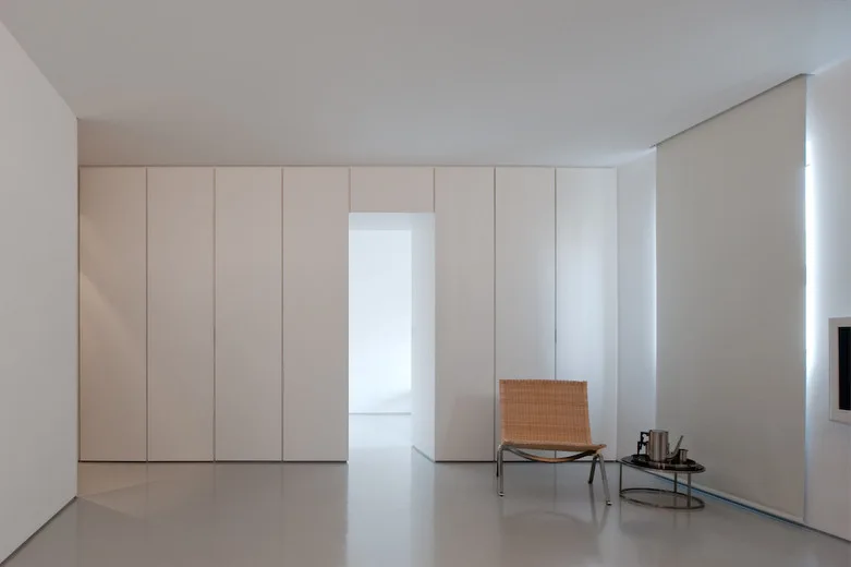 A minimalist room features seamless white built-in wall cabinets, a woven armchair, and a small metal side table on glossy light grey flooring.