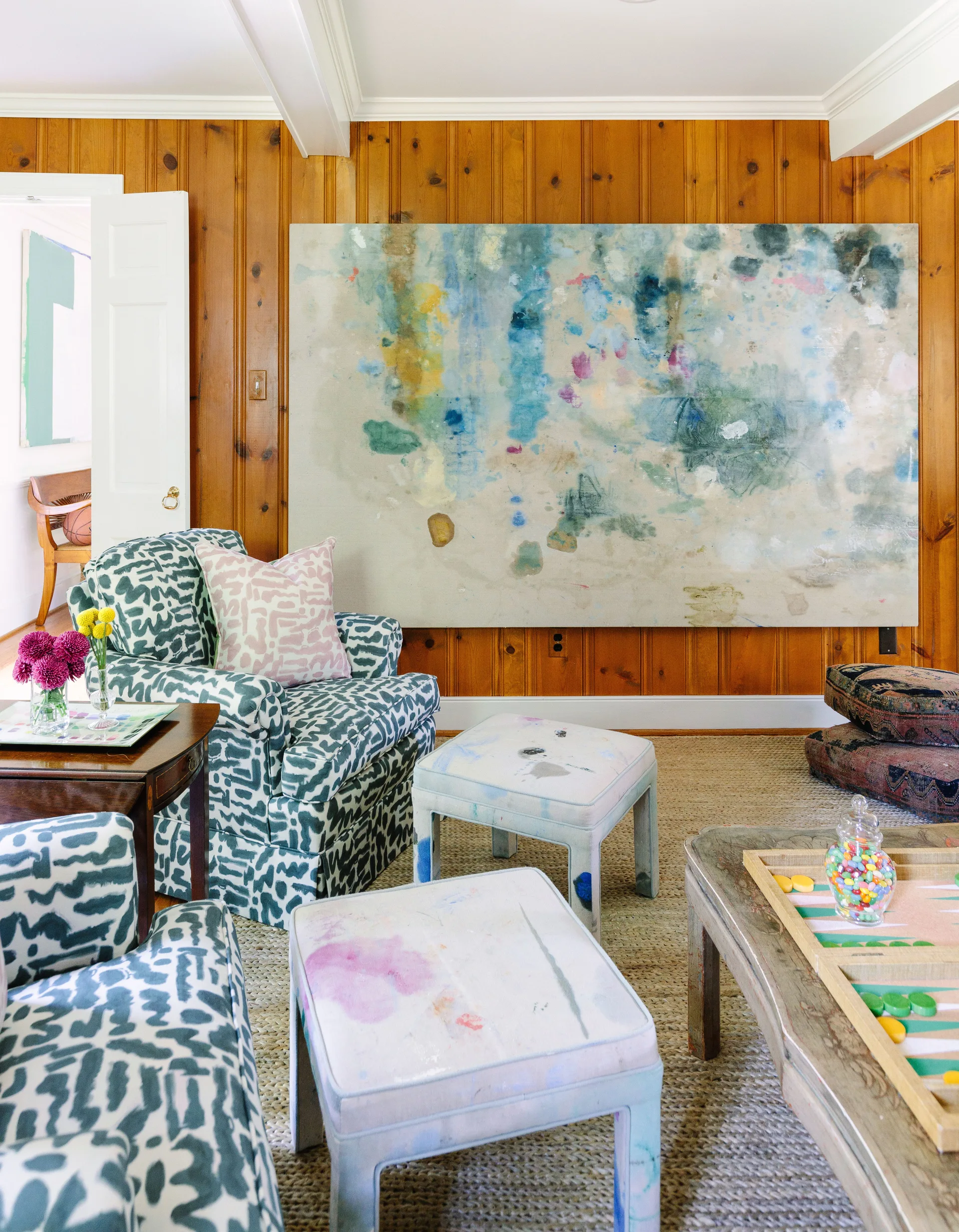A living room featuring vertical wood paneled walls, a large abstract painting, patterned blue and white armchairs, and two painted ottomans.