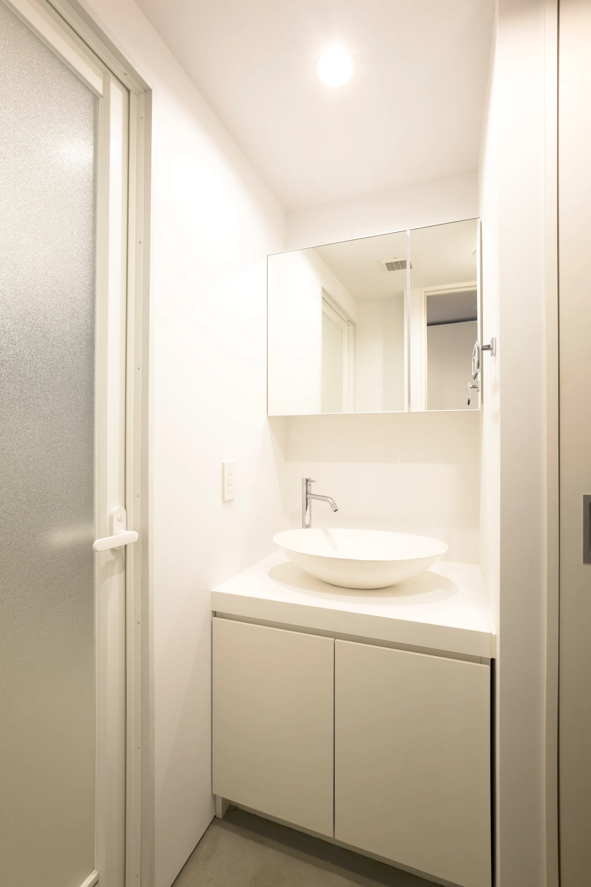 A minimalist white bathroom vanity features a ceramic vessel bowl sink, a chrome faucet, a mirrored medicine cabinet, and an adjacent frosted glass door.