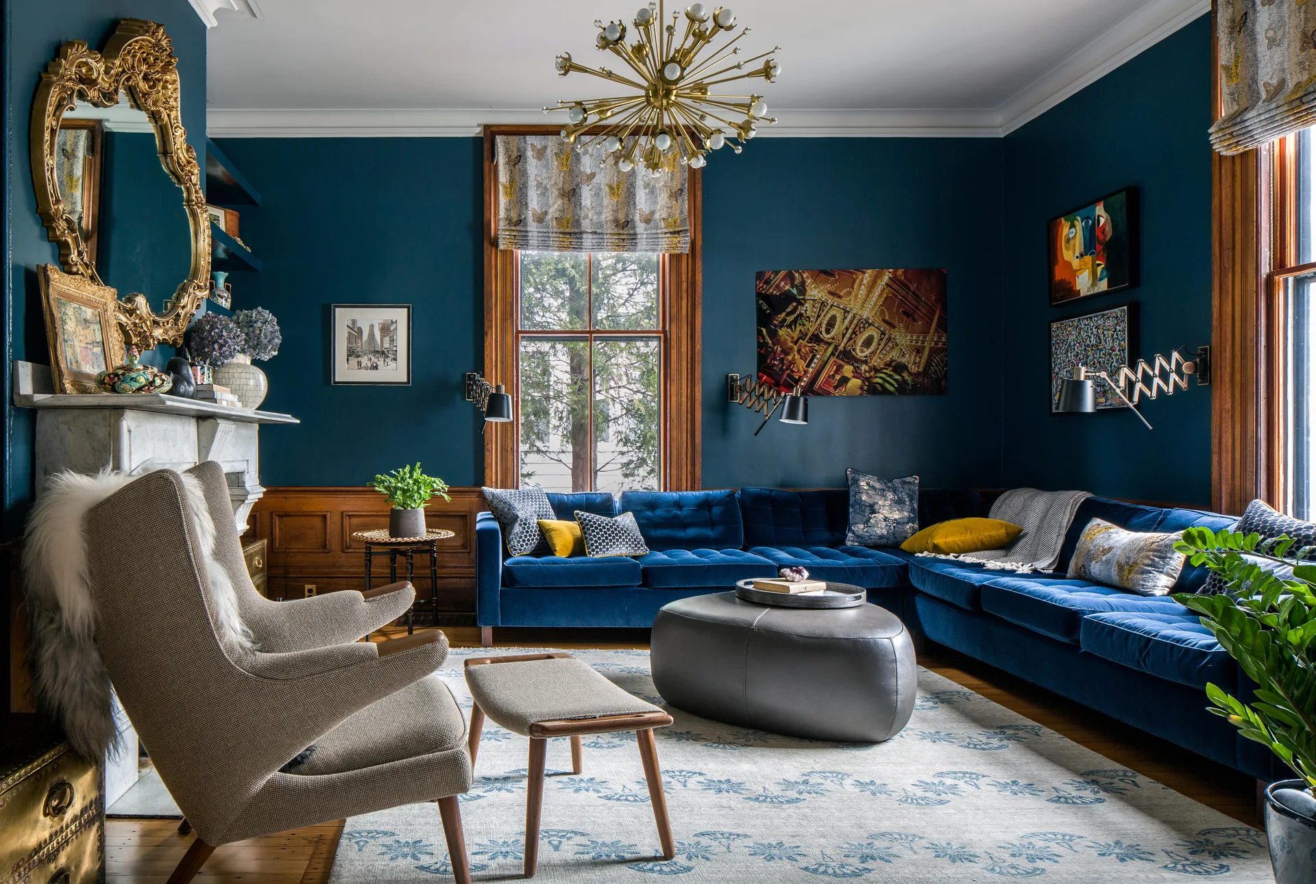 A living room features a deep blue velvet sectional sofa, dark blue walls with wood wainscoting, an ornate gold mirror, and a brass sputnik chandelier.